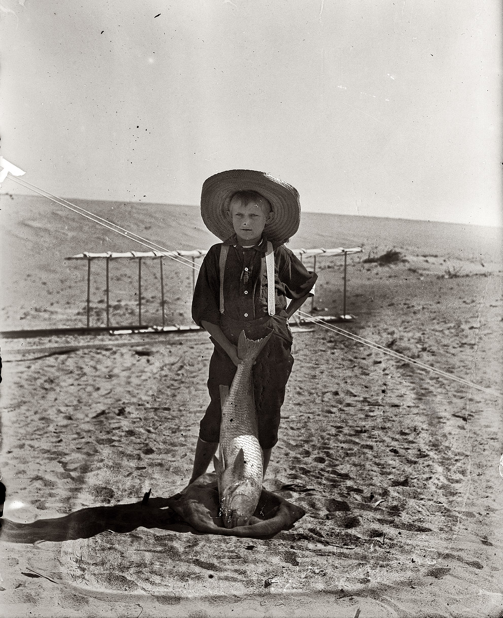 Tom Tate, the son of Captain Tate's half-brother Daniel Tate, poses with a drum fish in front of a 1900 Wright glider. Photograph by either Orville or Wilbur Wright, 1900. View full size.