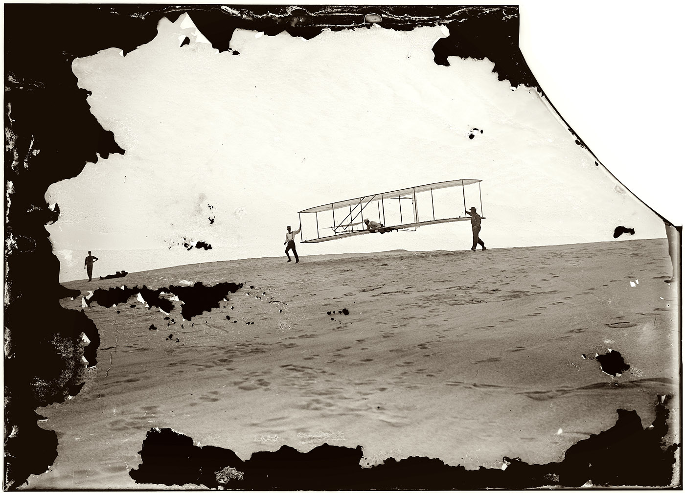 Kitty Hawk, North Carolina. "Start of a glide; Wilbur in motion at left holding one end of glider (rebuilt with single vertical rudder), Orville lying prone in machine, and Dan Tate at right." October 10, 1902. 5x7 dry-plate glass negative attributed to the Wright Brothers. View full size.