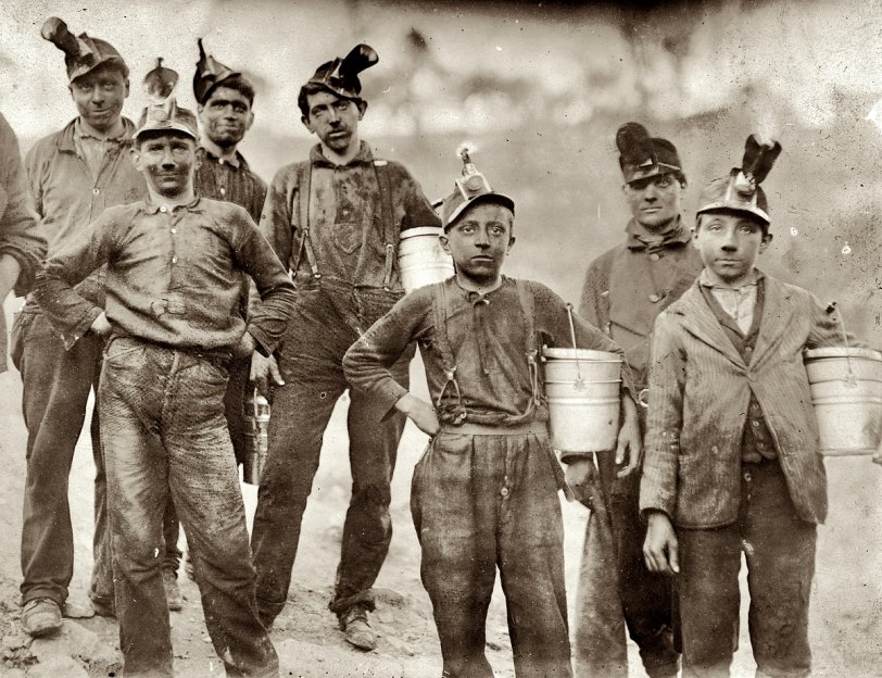 "Drivers and Trappers Going Home." Barnesville Mine, Fairmont, West Virginia. October 1908. View full size. Photograph by Lewis Wickes Hine. Note oil-wick lamps on the hats, and lunch pails. Trappers were boys who opened trapdoors to let coal cars pass and then closed them again to maintain proper airflow in the tunnel ventilation system.
One trapper's description of the job, which paid about $1.60 a day:
Trappers were responsible for opening and closing the underground  ventilation doors.  In those old mines, they had a system of doors between sections to direct the flow of air. Air was supposed to go up the main haulage and back to the fan.  So a trapper sat all day by his door with an oil lamp on his cap.  There was a "manhole" - a shelter hole in the wall by the track. The motorman would blink his light at me, and I'd throw the switch and open the door for him. Then, I'd jump into the manway until he was past, and run out and close the door. A trip would come along about every hour. Was I bored or lonely? Well, it was my job.
