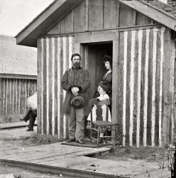 1865. "City Point, Virginia. Brig. Gen. John A. Rawlins, wife and child at Grant's headquarters." Wet plate glass negative, photographer unknown. View full size.
J.A.R.JOHN AARON RAWLINS was born in Galena, Illinois, on 13 February 1831; attended local schools followed by eighteen months at Rock River Seminary at Mount Morris, Illinois; studied law in the office of Isaac P. Stevens of Galena and was admitted to the bar in 1854; practiced law in partnership with Stevens and later with one of his own pupils, David Sheean; married his first wife, Emily Smith, 1856; was city attorney in 1857; was nominated a presidential elector on the Douglas ticket, 1860; helped organize the 45th Illinois Infantry and was designated a major in the regiment; was requested by Colonel Ulysses S. Grant of the 21st Illinois Infantry to accept a commission as lieutenant and assignment as Grant’s aide-de-camp; was appointed captain and assistant adjutant general of volunteers on Grant’s staff, 1861; lost his wife to tuberculosis; served as Grant’s principal adviser; was promoted to major in May 1862, lieutenant colonel in November 1862, and brigadier general of volunteers, August 1863; married Mary Hurlbut, 1863; was designated chief of staff of the Army, 1865; was brevetted major general of volunteers in February 1865 and of the regular army in April 1865; contracted tuberculosis; attempted to restore his health by accompanying Grenville Dodge on a survey of the proposed route of the Union Pacific Railroad to Salt Lake City; served as Secretary of War, 13 March 1869–6 September 1869; died in office in Washington, D.C., on 6 September 1869.
Hey, Mom, DadWhere's the rest of him?
WyomingIt should be noted that General Rawlins was U.S. Grant's closest associate during the Civil War and served as Secretary of War during Grant's presidency but only for a few months, dying of "consumption" in 1869. Rawlins, Wyoming, is named after him.
Press your pants?When was the crease in pant legs introduced?  This senior officer, a brigadier general, does not sport one in the mid 1860s, as did most men in dress pants. To our modern eye the men look unkempt.
Hirsute legal eagleHe may not look like much, but Rawlins rose to the rank of brevet major general and bragidier general in the regular army by the end, or just after, the Civil War.  His dates were 1831 to 1869.  
He was an attorney in civilian life, served US Grant as an adjutant general, and became Grant's first Secretary of War.
Photos of men of this era often carry with them the effect that the gentlemen pictured are elderly.  Here, in 1865, Rawlins is 33 or 34 years old.
US Grant was born in 1822. He was in his early 40s when he assumed the command of the Army of the Potomac.
To my eye, Robert E. Lee, who looks in his photos to have been in his 60s or 70s during the Civil War, was born in in 1807.  He was really just in his 50s when he was commanding the Army of Northern Virginia.  Lee was an age peer of President Lincoln (1809-1865), who was only 56 when he was assassinated.  But Lincoln always appeared much older, in my view.
Must be the beards.
Rank has its privilegesIf this is the General's quarters, can you imagine the enlisted men's?
What&#039;s that thing What's that thing that looks bolted on to the right of the porch, by the chair?
[A boot scraper. - Dave]
Ubiquitous Boot ScraperThat boot scraper was the first thing I noticed!  I can't help but think about the living conditions in what looks like a rough hut for a brig gen and his family.  And yet, there is a boot scraper!  Amazing.  I wonder if just the officers got them and was that one of the distinctions between their quarters and the quarters of the enlisted men -- to maintain some bit of genteel civility by scraping one's boots?
General RawlinsThe General was not a combat soldier but served as the principal trusted adviser to Grant. He was present when Lincoln appointed Grant command of the Army of the Potomac.
SmokerI do believe that is Grant himself around the corner puffing a cigar. He did tend to sit with legs crossed.Those cigars ended up killing him.
(The Gallery, Civil War)