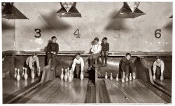 December 20, 1909. "Boys working in Arcade Bowling Alley, Trenton, New Jersey. Photo taken late at night. The boys work until midnight and later." Photograph and caption by Lewis Wickes Hine. View full size.
Interesting...mix of regular and duckpin bowling on adjacent lanes.
Denny Gill
Chugiak, Alaska
Duck pinsMy Dad told us kids that he once worked in a bowling alley setting duck pins. Didn't say how much he was paid or anything else about this job. This would have been in the 1930s in or around Clairton, PA, south of Pittsburgh. Later he would work as a hod carrier and parking cars in a parking garage before joining the Marines in 1940. He had a lot to say about these jobs.  
Joe Bartolini
West Columbia, SC 
PercussionThose duckpins look like upside down maracas.
Duck PinsDuck Pins is still a fairly popular variant of bowling in some areas. It's the preferred version in Quebec for example. Candle Pins (where the pins are basically straight tubes with only a slight bulge in the middle) is the found mostly in Eastern Canada and New England.
Concentrate!Those kids must have been very distracting to the bowler.
[I think they stayed behind the tarps until the ball came through. - Dave]
The Tarps *I wouldn't bet on the pin setters dropping behind the tarps.  Those are there to take most of the momentum from the balls so that they drop into the pit at the end of the alley. Needless to say the balls hit there with a pretty substantial force. Standing behind them and having limited visibility would be dangerous when some guy is hurling a 16 lb. bowling ball at you. As I understand it most later bowling alleys had a platform above the alley like the one these kids are sitting on but it had a sort of screen or wall in front of it. When a ball went through the kids would drop down set the pins and jump back up all while hoping that some sadistic SOB wasn't throwing another ball to hit them.
[There are a few photos of the kids coming out from behind the tarps, which are not fastened at the bottom. - Dave]
(The Gallery, Kids, Lewis Hine, Sports)
