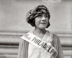 &nbsp; &nbsp; &nbsp; &nbsp; UPDATE: This is the lovely Miss Nellie Orr!
Washington, D.C., circa 1921. "Nellie [Illegible], Miss Philadelphia." Perhaps someone out there can put a last name to this winsome face. View full size.
Like some kind of sea creatureWhat a fantastically weird hat.
Nellie OrrI think it was Orr.  
http://www.misspa.org/past.htm
Up in Here"Why are ya'all up in my grill?"
Actually,she's the cutest beauty pageant contestant ever. 
Miss Nellie OrrMiss Nellie Orr, Miss Philly 1921 and one of only eight contestants in the first (1921) Miss America pageant in Atlantic City.
http://www.pageantopolis.com/international/America_1920.htm
Nellie looked "spunky".
Miss Nellie OrrThe Coshocton Tribune (Ohio), 10 September 1921 (via NewspaperArchive.com):
SHE'LL REPRESENT MISS PHILADELPHIA IN BEAUTY REVIEW
Miss Nellie Orr of Philadelphia was chosen in the recent beauty contest to represent the Quaker City in the beauty review to be held in Atlantic City some time in September.
Literally winsomeNellie didn't have much equipment even by the standards of that day (teeth didn't matter, shape did!) but something in her look tells me she would have been a formidable competitor in anything she chose.  She certainly didn't get the scarred lip and broken tooth from tea parties or knitting.
Miss Illegible: 1921The girl was Nellie Orr. See this link for list of Miss Philadelphia winners.
http://www.misspa.org/past.htm 
Whoa NellieMISS AMERICA 1921
1921 September 7
8 entries
Result
1  WASHINGTON DC - Margaret Gorman
Remainder
CAMDEN (NJ) - Kathryn M. Gearon
HARRISBURG (PA) - Emma Pharo
NEW YORK CITY (NY) - Virginia Lee
NEWARK (NJ) - Margaret Bates
OCEAN CITY (NJ) - Hazel Harris
PHILADELPHIA (PA) - Nellie Orr
PITTSBURGH (PA) - Thelma Matthews
Miss Orr: 1921Looks to the future and tells herself: "I'm gonna be the best Miss Philadelphia ever and with my winnings I'm gonna buy another letter or two for my pathetically short last name!"
Near Miss In 1921 Nellie Orr competed as Miss Philly in what would soon become known as the Miss America Pageant, where out of 500 contestants in the "bathers' review," she finished second. Something must have been stuffed -- either the ballot box, or ... 
Heeere she isIn the Racine Journal-News, same great hat
Poor PhiladelphiaFlat as a pancake, foul teeth! My God! I wonder what her contenders looked like.
Prosthodontically speakingMiss Orr seems to be sporting a none too artfully fashioned porcelain jacket crown. Or is it an inlay?
Her nameYes it was Orr, and she was my great-grandmother on my mom's side. From what my grandfather has told me about her, she was very spunky and outspoken. He used to tell me I reminded him of her! 
Nellie OrrNellie was my mother's older sister.  My mother is 82 and still lives in Haddon Heights, NJ.
I hope Heaven is far awayOtherwise, she's still embarrassed every time someone looks at this picture. She's probably saying something on the order of, "Of all the photos taken of me, how did this get to be the one people are still looking at? Now that it is on Shorpy, I will never live it down!"
Equipped Just FineMiss Nellie is actually built perfectly for the standards of the day. By 1921 the flapper era was in full swing, emphasizing an almost boyish look with bobbed hair, flattened breasts and few visible curves. It was a reaction to the Victorian style of very long hair and fairly extreme curves accentuated by a corset. It's no surprise she would finish second in the "bathers' revue."
(Full disclosure: my grandmother was a flapper. Her hair had never been cut until 1919 at age 12, when she got a bob. She told me her father didn't speak to her for weeks!)
What Happened to Miss Philadelphia 1921 Nellie Orr?Does anyone know what became of Nellie Orr? I am researching all eight of the 1921 Miss America Contestants from the first contest and located info on all except for Nellie.  Looking for her parents names, Nellie’s married name and when she passed away.    Many thanks!  You can contact me at  NRFB59@aol.com
Nellie Orr at Miss America 1921Here she is in her black taffeta swimsuit.

(The Gallery, D.C., Natl Photo)