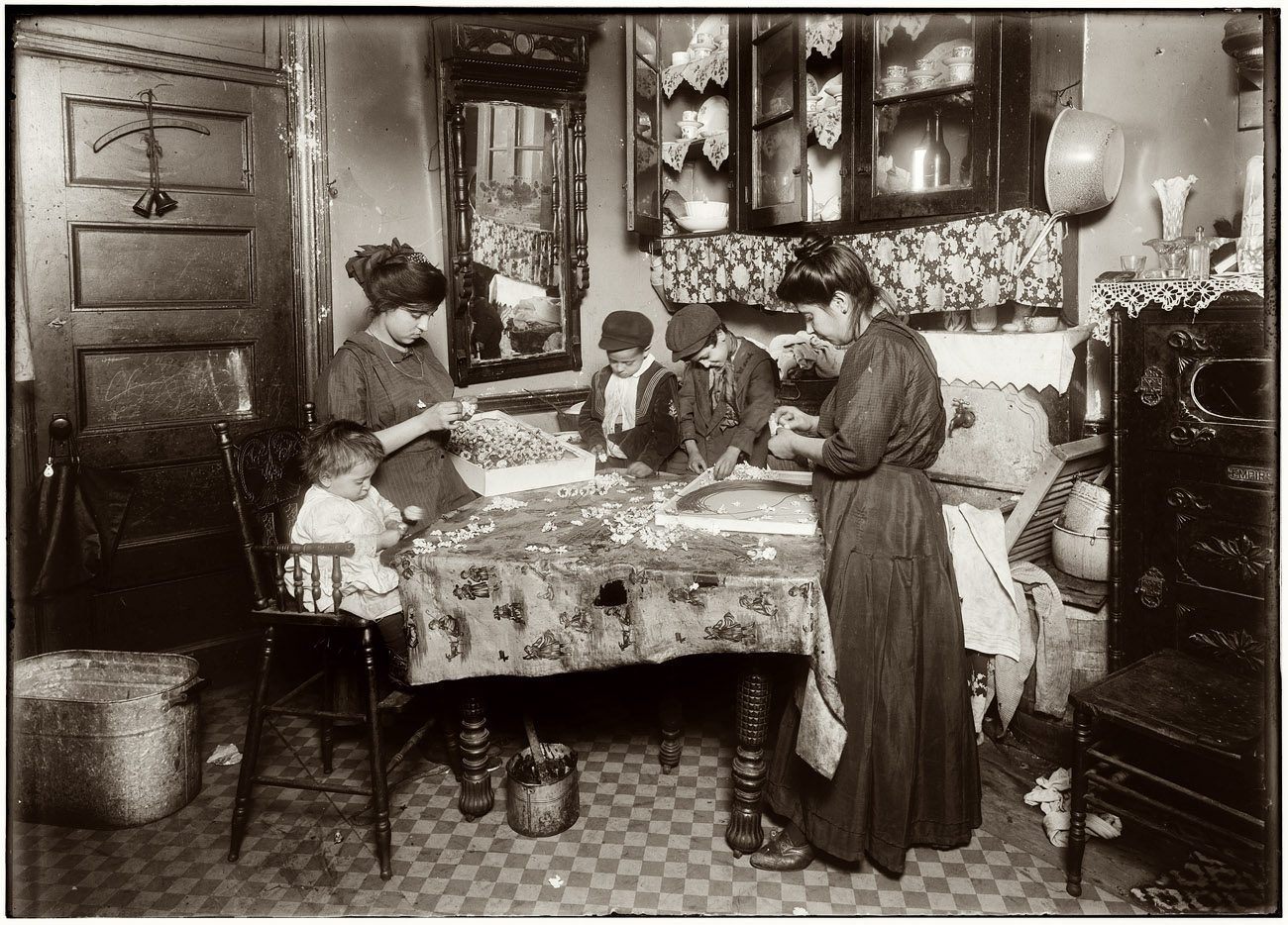 December 1911. Family of Mrs. Mette making flowers in a very dirty tenement, 302 Mott Street, top floor. Josephine, 13, helps outside school hours until 9 P.M. sometimes. She is soon to be 14 and expects to go to work in an embroidery factory. Says she worked in that factory all last summer. Nicholas, 6 years old and Johnnie, 8 yrs. The old work some. All together earn only 40 to 50 cents a day. Baby (20 months old) plays with the flowers, and they expect he can help a little before long. The father drives a coach (or hack) irregularly. View full size. Photo and caption by Lewis Wickes Hine.