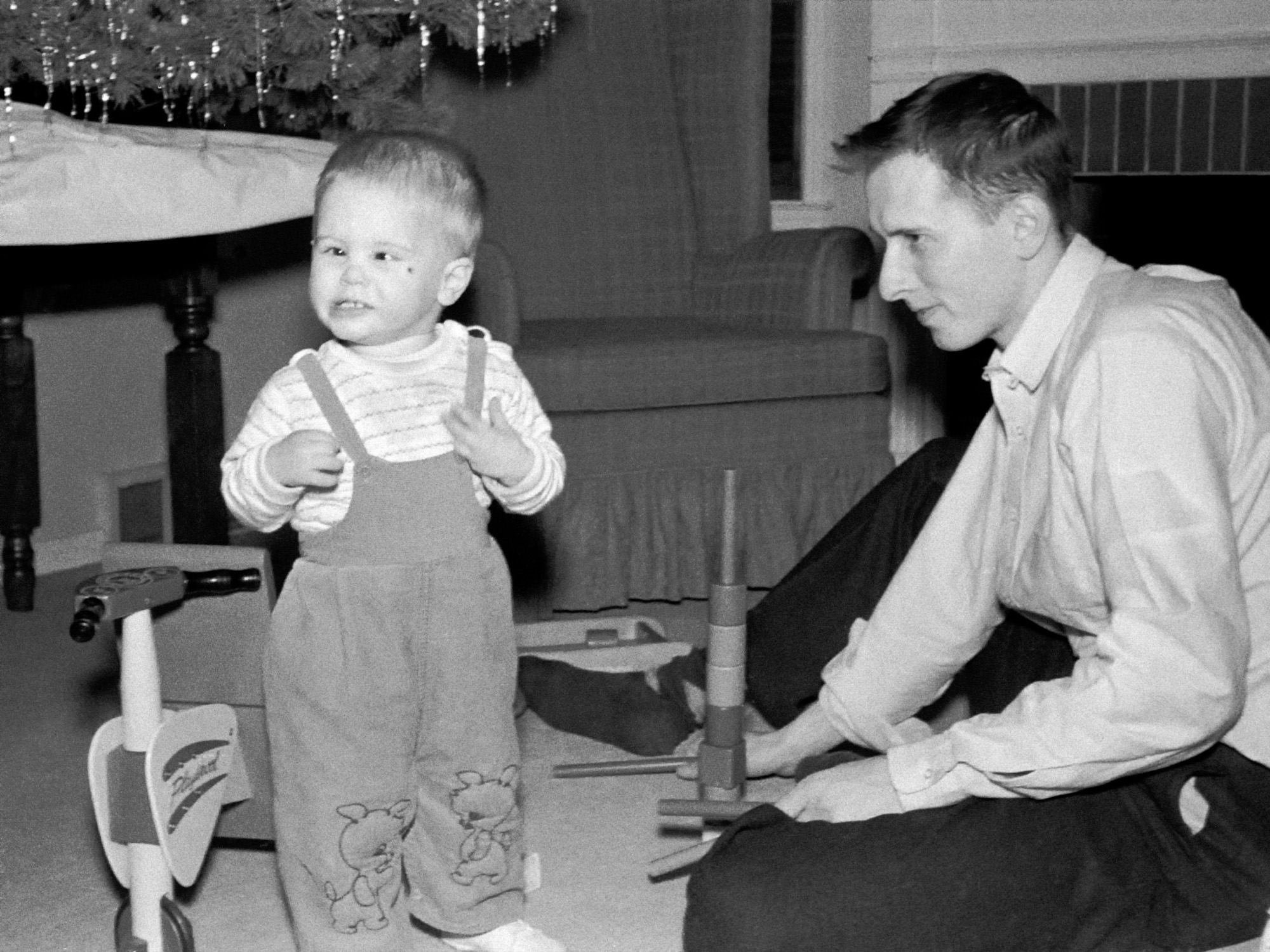 This is me celebrating my second Christmas.  While Mom handles the camera (a rarity) Dad (29-years-old) and I are in the front room of our first house on the North East side of Indianapolis.

The bump by my left eye has nothing to do with the left eye's inward turn. My congenital esotropia was corrected via surgery about a year later.

The Playschool trike lasted many years through multiple siblings but is with us no more.  The flat wagon behind Dad's right foot holds the various blocks and rods that we are playing with. My Mom still has the blocks and wheeled wagon - now used by a second generation. View full size.