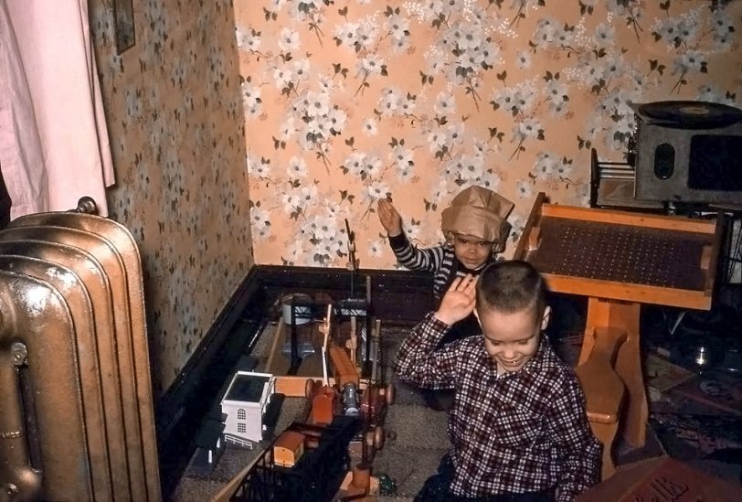 Here is a picture of younger brother George, and I, playing with our "shared" American Flyer c.1958. The "steam" locomotive and tender cannot be seen, but on the floor you may spot several familiar items: signal bridge, telephone pole, water tower, small trackside tool shed. There are two windmills made of tinker toys, too. We had a cow that would block the track and quickly move out of the way when the train approached. Also in the room, with fanciful wallpaper, note the steam radiator, the peg desk and the record player. Burl Ives' "Bimbo" is probably playing. What imagination! What fun! 
Kodachrome slide taken in Clinton, Iowa, c.1958. View full size.
