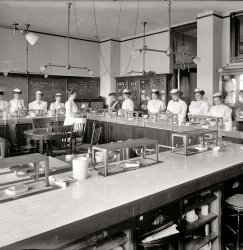 "McKinley School lab." Home economics at McKinley High School in Washington circa 1910. Harris &amp; Ewing Collection glass negative. View full size.
Asbestos mat?Well at least they did not have guns, drugs, or knives. 
Ok, I&#039;ll biteWhat are the twin chains hanging from the ceiling for? 
[My guess would be damper chains. - Dave]
Oh GoodIt's comforting to see that "1 - asbestos mat" is included in the chalkboard list of kitchen must-haves!
Chains on the ceilingIf it's anything like the high school I went to the chains operate the sashes on skylights. 
As far as the asbestos mat,As far as the asbestos mat, Asbestos has a bad reputation because of asbestosis, but a well made asbestos blanket is perfectly safe as long as you don't let it get frayed.
However, I think Dave's got the title wrong on this one... these young ladies aren't studying to be housewives, they're studying to be the hired help, kitchen maids or cooks, judging by how they're dressed.  The room isn't even set up right to properly emulate a "normal" home of the era.
Of course, I could be wrong....
[McKinley Manual Training School was, at least in name, a vocational high school. But that doesn't mean these ladies aren't learning the domestic arts while they're in the cooking lab. The aprons, like the aprons in any lab or kitchen, are there to protect your clothes. Something tells me there weren't many white girls in circa 1910 Washington pursuing careers in domestic service. - Dave]
Combo lightsIt appears those lights are combination gas and electric- with a gas jet pointing up behind each bulb.
[They're gas fixtures that have been wired for electricity. A common practice when electricity was replacing gas for illumination. - Dave]
All right Dave, explainSo why do you think there weren't many white girls in circa 1910 Washington pursuing careers in domestic service? Seriously, do you know something I don't? From what I've read, photographs, etc, there seem to have been scads of white servants in that era. It's pretty darn common vocation for a girl of modest means, and those frilly white caps indicate a servant uniform. You don't see many housewives wearing those - strictly for the help. I'm not sure how being in Washington in 1910 would have changed that. Was there a highly localized white-domestic plague that year?
[Anything is possible. Maybe McKinley High did have a "maid training" program where you got to play dress-up. Most of the girls shown taking food-preparation instruction in photos from this era are shown wearing aprons and caps. Then as now, food preparers in an institutional setting would wear a cap or hairnet. If these girls had career aspirations that involved cooking, I think nursing or restaurant work would be the more likely choices. The McKinley course offerings as listed in the Washington Post archives are pretty general -- machine shop, carpentry, motor repair, home economics. The classroom in our photo was the "domestic science" (home economics) lab. Instruction was given in "plain and fancy cookery," invalid cookery and menu planning. - Dave]

I don&#039;t think it&#039;s jettingThe light fixture on the right has tubing coming out of what would be the jets, so I'm not sure where the flame was supposed to be. Unless... the ends were cut off and fixed with lightbulbs. But they're sorta facing downward instead of upward.
[That's a rubber hose for something like a Bunsen burner. So the gas must still be connected. - Dave]
I would have failed the classSince I really hate to cook! 
Gas hoses for tabletop useScary as it seems to me, combination gas and electric chandeliers also provided convenient sources for portable gas table fixtures via rubber hoses such as the one in this photo. I've seen a 1910 photo of a gas table lamp in the parlor of Ulysses S. Grant Jr.'s house in San Diego, using just such a hose dangling down from a gascock on the chandelier overhead.
Wires and Gas When I first moved into my old house in Atlanta, a house built in 1910 or so, I found cloth-insulated wires for overhead lamps running through the gaspipes to every room ceiling.  It confused me until I realized that electricity was added onto the house in the twenties.  There were also the "knob and tube" ceramic insulators all over the attic.  I got that overhauled quick!
(The Gallery, D.C., Education, Schools, Harris + Ewing, Kitchens etc.)