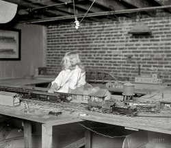 December 11, 1925. Washington, D.C. "Margaret Swartzell -- Swartzell railroad system." Not just a model train, it's a "system" -- who can tell us more? National Photo Company Collection glass negative. View full size.
A frustrated engineer!According to that 1934 Popular Science article, Mr. Swartzell had attended the railroad engineering program at the U of I, Urbana-Champaign, which was one of the two most respected programs in steam locomotive design at the time (the other was Purdue--you know, the "Boilermakers").  Mr. Swartzell, it seems, had real talent.  I'm sure he made excellent money in his father's real estate business, but technology must have been a hard thing to give up.  Especially such a romantic technology--the call of a steam whistle still tugs at our heartstrings to this day.  
I suppose it was for the best, though.  Steam was essentially dead within 25 years of this photo.
A practical treatiseEarly model railroaders used mechanisms from England. English models were built to 7mm = 1 foot (1:43). When the Americans compared the size of locomotives it was thought that ¼ inch = 1 foot (1:48) was close enough. The boilers were made of lead pipe solder (origin of the phrase "lead pipe modelers"). Details were cast in homemade patterns. Tin cans and old crates were used for various parts. The commercial model kits were very expensive. Railroads had their apprentices build working models of steam engines in larger scales for practice. Lindsay Publications Inc. has old books on this subject.   
Railroad Real EstateFrom Popular Science, Oct. 1934.

GrungyDidn't they ever clean anything back then? Maybe they were going for an authentic rail yard look. What's that hanging from the spider web under the table? Bug? Leaf? Booger?
The system must be larger than what we see here; looks like it goes through a tunnel in the wall in that back corner.
The Man&#039;s domainI doubt very much that Mrs. Swartzell ventured down in the basement (or out to the garage, just as likely) to dust the train set.
Our basements over the years were cement or dirt floors where spiders and other buggly critters abounded. Obviously Mr. Swartzell cared little about dusting the odd footprint off the platform (probably had to plug and unplug from the ceiling light every time he wanted to use the trains). He probably had more interest in playing with his trains than worrying about whether or not the odd cocoon or spider eggball hung from the bottom of his tracks.
J.N. Swartzell...and from Popular Mechanics, 1925
PiffleIt's not as exaborate as the basement railway of two friends in the 50s whose father worked for Lionel.
I coveted the GG1, and the sound of trains going over the maze of switches in the rail yard they parked in.
Amazing model railroad for the time.What is most amazing about this model railroad is that it is two rail at a time when toy trains like Lionel and American Flyer were three rail.  The effort that went into insulating all the wheels on all the locomotives and rolling stock is mind boggling since all the modern plastics and adhesives we have today were unknown and not available.  The two major model railroad magazines, "Model Railroad Craftsman" and "Model Railoroader" go back to the early 1930's so there were not a lot of resources for Mr. Swartzell to refer to.
&quot;Every Bit of the System Hand-Built!&quot;If the Popular Science article is correct and this fellow built everything in the photographs himself, by hand, that's not a "piffling" achievement. It's an accomplishment that deserved every bit of recognition he received, in my opinion.
Eddie LaytonA friend of mine who passed away a few years ago, Eddie Layton, was the organist at Yankee Stadium. He had a model railroad collection that he assembled over many years. It ran on a reinforced plywood panel about 12 feet by 10 feet. He lived in an apartment in Forest hills, Queens. He had it in his living room, rigged to lines that he could lower from the ceiling to the floor. Eddie was the subject of a well known Trivia question, "Who was the man that played for the Yankees, Knicks and Rangers in the same season?" The answer was of course Eddie, as he was also the organist at Madison Square Garden. Ironically he also  played for the NY Islanders at the Nassau Coliseum  for a few of those seasons as well.
Fair River JunctionMr. Swartzell's layout was also featured in a 1929 article in  Machinists Monthly Journal, the "Official Organ of the International Association of Machinists."  Fortunately, that issue is available in PDF format courtesy of Georgia State University Library. (link to PDF [3.2 MB], article begins on pg. 584.)  The photos in that article, poorly rendered within the PDF, are also in the LOC archives. Perhaps Dave will share them with us one day.
A few excerpts...

The "railroad" is supposed to be located in a valley of the Alleghenys, with the mountains away to the west and north. The main line stretches westward, enters a tunnel, swings around a long loop and returns, down the banks of a river, across a steel bridge, to the terminal.
In the west foreground is the roundhouse, with turntable, coal dock, oil, sand and supply house, etc. Behind it is the back shop, with two "drop-pits" for light repairs and, beside it, the freight and storage yards.
"To the east is the coach yard and the express and freight depot while the main switch tower and dispatcher's office are opposite the passenger depot, with a maze of switches and crossovers between.
The town lies beyond the main line, on the river flats, with hotel, farm houses, residences, etc. Highways run across the flats and up into the mountains.
Everything is accurately built to a scale of one-fourth of an inch to the foot.
...
"It is not, as has been stated, a reproduction of the B. &amp; O. Mountain Division," Mr. Swartzell said. "It is, however, a faithful copy of B. &amp; O. equipment located at an imaginary mountain division point which I have called 'Fair River.'"
...
The passenger rolling stock consists of Pullmans, day coaches, observation and chair cars, baggage, mail, express and express refrigerators and even combination mail, baggage and express cars.
The freight equipment is equally varied, but much of it is out of date and must be replaced when, as Superintendent Swartzell says, "the appropriations for maintenance of equipment permit."
...
"That tunnel is a problem," Swartzell confided. "It is right at the foot of a steep grade with sharp curves. "The worst wreck the division ever had occurred right inside it. You have recently written something about freak wrecks. This was a queer one.
"We sent out a solid express and baggage train and a freight right behind it, westbound. A careless baggageman left a door open and some trunks fell out on the opposite main. The freight had been swung over on the east-bound main so it hit the trunks and piled up. We had a lot of trouble picking up the wreckage and clearing the line."


Sidenote: This basement looks so typical of the row-homes in D.C: exposed brick walls and beams spanning the width of the house. One of the first things that caught my eye was the brickwork: another fine vernacular sample of "American" or "Common" bond.
Fascinating!The little Girl couldn't have been more perfect for this photo! Her expression is priceless. Then there's the detail in the buildings, cars and engines. The engineer who designed this layout had the passion! If one looks under the left half of the table, one clearly sees whatlooks to be left over track. And the water tower! Very nice. There's quite a bit going on here. 
CellargatorMy great-aunts bought a tiny alligator back from their jaunt to Florida in the 1920s. Back then, those living souvenirs were all the rage. After a few months, it disappeared from its tank. They figured it would show up dessicated under a radiator within a few months.
Three years later, one of them went down to shovel coal for the stove. She heard a loud hissing and saw red eyes glowing down in a corner underneath the foundation, behind the coal cellar.
They got the fire department and the police to kill the "monster," which was now about three feet long. It had dug itself a nice warm wet hole in the dirt floor, where it survived eating rats and stray cats and squirrels.
The hide was nailed to the garage, where it still freaked me out 40 years later.
End of the LineWashington Post, Nov. 20, 1937
J.N. Swartzell's Funeral Is Set for Tomorrow
Funeral services for John N. Swartzell, retired Washington business man, will be held at 11 a. m. tomorrow at his home, 2725 Thirty-sixth place northwest. Burial will be in Rock Creek Cemetery.
Swartzell, who was 47-years-old, died Thursday at his home. He retired in 1925 from the firm of Swartzell, Rheem &amp; Hensey because of ill health. His father, G. W. F. Swartzell, was one of the founders of the firm, which failed in 1931.
Born in Washington, Swartzell was educated at Friends School and at George Washington University, where he was a member of Theta Delta Chi.
He was a past president and honorary member of the Civitan Club, secretary of the board of managers of the Methodist Home and a director of the Columbia National Bank. He was also a past master of Temple Noyes Masonic Lodge and a member of Mount Pleasant Chapter, Royal Arch Masons.
Surviving are his wife, Mrs. Anna Drury Swartzell; a daughter, Margaret Swartzell; a sister, Mrs. C. C. Davis, and a brother, Henry R. Swartzell. All live in Washington.
B&amp;O Jr. According to the July 1936 issue of Model Railroader the name of the railroad was "The B&amp;O Jr." The article notes that Mr. Swartzell began construction of his model railroad "shortly after the end of the war."
(The Gallery, D.C., Kids, Natl Photo, Railroads)