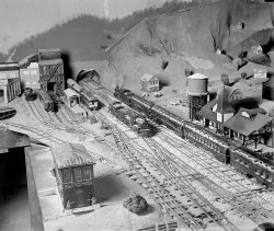 August 5, 1929. Washington, D.C. "Miniature R.R. of John N. Swartzell." National Photo Company Collection glass negative. View full size.
Miniature weatherScale model snow on the railroad cars and locomotives. Neat. Actually, would snow adhere to the main part of a steam loco?
ImpressiveI'm duly impressed with Mr. Swartzell's modeling skills, in 1929 he couldn't purchase much (if any) scale equipment. About all he purchased commercially in this photograph is the rail.
Looks Handmade     This pic shows attention to detail that is amazing. Whats more impressive is that this train set looks like it predates the trains we had in the 50s and 60s. The engines and cars look to be handmade out of wood or something. I am impressed. Is that supposed to be snow or sand on some of the cars?
Nice, butIt's missing stray dogs, a baseball game and roaming chickens.  
ScaleWow!  There are things on this layout that rival work of the great modelers today.  
One item is the forced perspective; where you model a building smaller than the rest of the structures so that it looks like it is in the distance.
Does Mr. Swartzell&#039;s layout still exist?Model train villages are such intense labors of love, but they can take up a lot of space. Was this one kept intact, perhaps transferred to another location? Or was it taken apart and stored, forgotten, in someone's attic as "Grandpa's Train Set"?
If you live on the East Coast and like model train villages, there is none better than "Roadside America," in Shartlesville, PA. (Route 78 between Allentown and Harrisburg.) From its description, you might dismiss it as corny hokum - and there is a bit of the cornball about it. But it truly is amazing. And it, like Mr. Swartzell's village, is the work of one man, essentially.
Their wives always knew where to find them.
For some amazingly accurate photos of Roadside America, try these.
Third railThe third rail is visible in this picture. Wherever there are gaps in the switches there are buttons or nails sticking up.. Early tinplaters modified three rail tracks like this. Another development was the outside third rail.
Three rail has the advantage of not having to insulate the
the frogs of the switches as two rail does. It also makes signaling simple. Two rail became popular as model trains became more detailed. 
No White Shoes, No ServiceNotice the men at the right with dark shoes. Cannot sit with the other patrons!
[It's amazing what you can see here, isn't it? - Dave]
FantasticAs someone in the world of model railroading (I'm a 3 railer if anyone is curious) it is always enjoyable to see what the pioneers of the hobby were able to accomplish with so little to work with.
Odds also are that the switches and trackage were  handlaid, and quite possibly much of the rolling stock and buildings were built from scratch.
Hand laid railThis is no "out of the box" rail layout. The ties are all individually set and the rail laid and spiked. The switches and frogs are all handmade and filed. Amazing track work, done only by the most smitten of model R.R.'ers. 
AuthenticityThat layout is filthy. Just like actual areas adjoining train tracks.
Timeless!It must have been even more impressive in color! Although today's scale material is more sophisticated, this man's work proves that imagination and hard work was and still is the basis to this "hobby." It's fun going in to the enlarged versions and looking at the detail that went into creating this layout. 
(The Gallery, D.C., Natl Photo, Railroads, Small Towns)