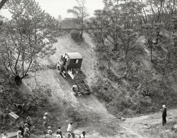 Washington, D.C., or vicinity circa 1928. "Four Wheel Drive hill-climbing demonstration." Our second look at this exciting test of trucks and traction. National Photo Company Collection glass negative. View full size.
This looks likeA stretch of Rock Creek Parkway between M and P Streets (before the bike path was paved).
MechanismIt looks like those rollers would be engaged against the rear wheels, in turn operating the exposed gears.  To lift a dump bed, perhaps?
Good LordSubtitled "Accident waiting to happen." 
Toothsome Winch! Wonder what the wicked looking winch on the back was to be used for. Pretty serious towing or dragging by the look of it. Note the rear towing hooks on the frame as well. 
FWD/Seagrave is still alive and well in Clintonville, Wisconsin, where the High School team is the “Truckers.” 
PTOIt looks like a Power Take Off unit driven by the drive shaft.  There would be a selector that switches between the axles and the PTO. 
The two large drums behind the rear wheels would accept a drive belt from some other piece of equipment and operated while the truck is stationary.
My grandfather had a similar one-drum arrangement on an old tractor from the '40s that was used to run a conveyor-elevator that went up into the hayloft of his barns.
LandmarksIt might be possible to figure out where this photo was taken if anyone recognizes the water tower[?] in the background above the truck. It looks like there may be a sign painted on it, but I can't make it out.
Hill climbing tipsWhether he makes it to the top depends a lot on how fast he was going when he started the climb. 
Rocks OnNotice the wooden box o' rocks and other stuff, including a shovel, behind the cab temporarily (judging by its cobbled-together look) mounted as far forward as possible in an effort to get some extra weight on those front driving wheels OR to minimize the possibility of a crowd-pleasing 1928 FWD truck wheelie. I wonder what water tower that is in the distance? 
One final thing, whatever the photographer was standing on, it's much higher than the far side of the draw/arroyo/small canyon. His position so near the action and unobscured view seem to indicate he was shooting from a building that stood on the edge on the high ground. This could have been at the Army's Aberdeen Proving Grounds in Maryland, not far from me, which has been testing military equipment including tanks and trucks since 1917. Note the uniformed gent in the foreground. At first I thought that was a boy with the bike, suggesting some informal setting open to anybody, but I think he's an adult, so it could be on government property. 
Kids to the front!Interesting that the kids and the one on the bike got in front of the suits and the Army officer observing the demo!
Disturbingly CloseThe spectators seem to be dangerously close to the action. In particular, the lone guy on the right seems vulnerable. I see no chains to safely hold this vehicle should it slip backwards, so why would one assume that it would fall back in a straight line? 
Friction winch rollersThose rollers are used for friction winches, a rope is wrapped around the roller several times, by pulling on the rope it grabs onto the roller like a friction clutch and transmits the power from the winch, easy way to control operation and loads of pulling power, the tighter you pull the rope the more power transmitted, let up on the rope and the pulling stops. The belt drive mentioned by PTO would not have had sides on the pulley; the pulley would be slightly crowned in the center but no sides or flanges.
(The Gallery, Cars, Trucks, Buses, D.C., Natl Photo)