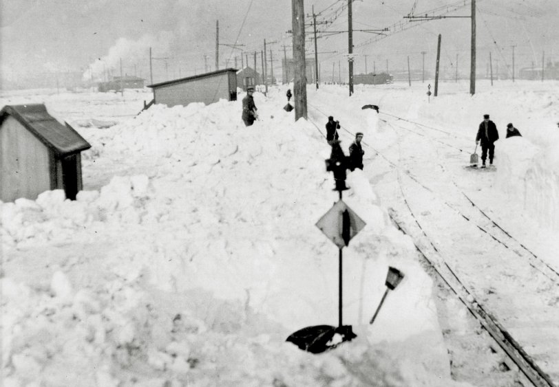 East Chicago, Indiana, January, 1918. The Chicago, Lake Shore and South Bend Interurban Railroad. Volunteers and railroad employees are trying to clear snow from a crossover located near the junction where streetcars and interurbans shared track. The South Shore ran on the principal east-west street, Chicago Avenue.  Along with South Bend to Chicago trains, a 3.4-mile streetcar line to the Indiana Harbor section of town was operated, primarily for shift-change steelworkers and a connection with the Gary Railways line at Michigan and Guthrie Street.
In the background, equipment can be seen hopelessly stuck on the streetcar track.  It was critical to free the line, as milk and other essentials were starting to run out.
(C. Edward Hedstrom Collection photo - given to me in 1985) View full size.

