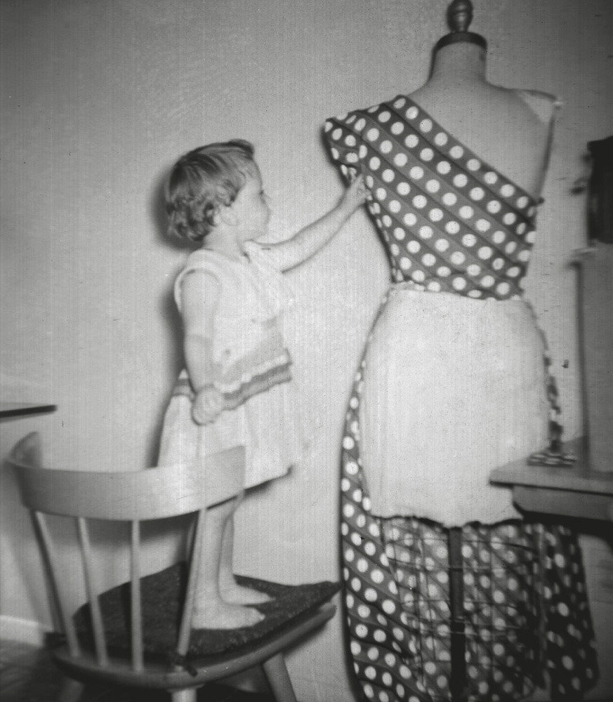 My mother’s whole life was (and to an extent still is) seen through a lens of apparel. Her mother was a milliner (made women’s hats during the first quarter of the 20th century, when everybody wore hats). Her father was a pattern-maker. My mother graduated from FIT (Fashion Institute of Technology) in New York City. She designed and made her clothes. She designed and made my clothes. She never bought a curtain, bedspread, or furniture slip cover in her life. She made them. If you were looking for her in the house, she would not be doing heavy duty cleaning. We had hired help for that. But she might be making fashion sketches, or sitting at the sewing machine.

Here, three-year-old me stands on a seat cushion she made, which is on one of our Paul McCobb mid-century modern chairs, to examine to polka dot fabric she has draped on her dress form, as she starts the pattern-making process for another dress. 