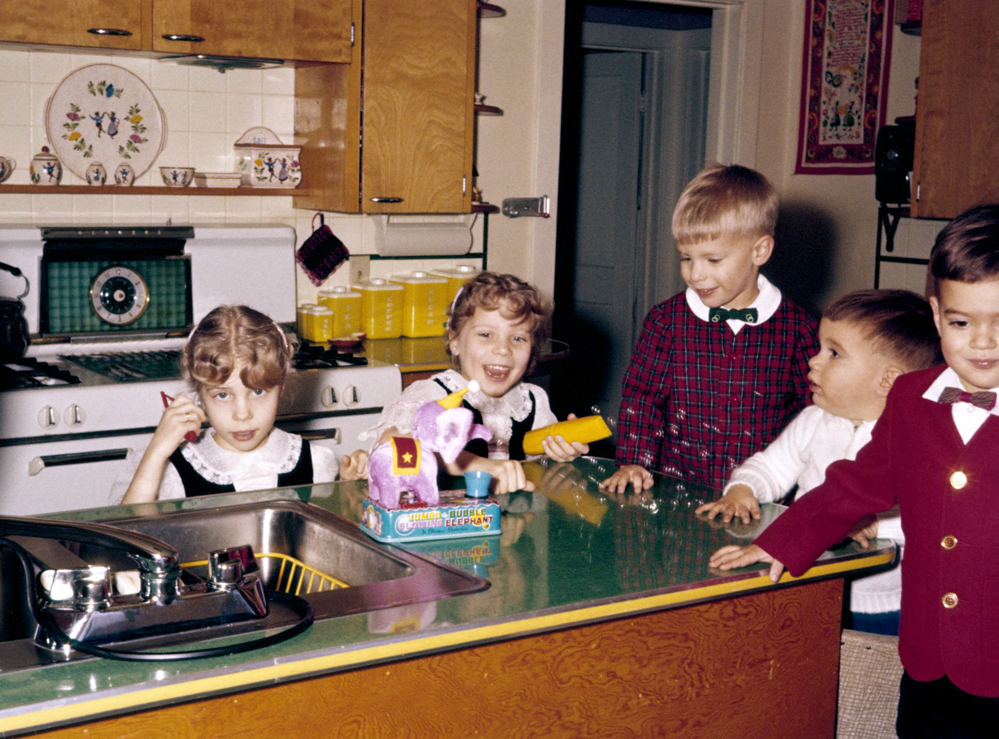 My brother's third birthday, this slide an Ektachrome (E-2 process in 1959?), East Aurora, NY, again taken with my father's Minolta A rangefinder. The cousins seem to be wearing exactly what they wore to last year's party. Didn't they grow?? I (in white) put in my appearance for the first time this year. At the center of attention (for at least a few of us) is Jumbo the Bubble Blowing Elephant. You can see that my mother's favorite color is yellow, and the folksy/folkdance theme continues in our kitchen. Potholders. Swing-Away can opener. Farberware tea kettle. Green dial wall-phone (I remember our number started with NL-2). Bow-ties. I've also uploaded a picture from his first birthday. View full size.