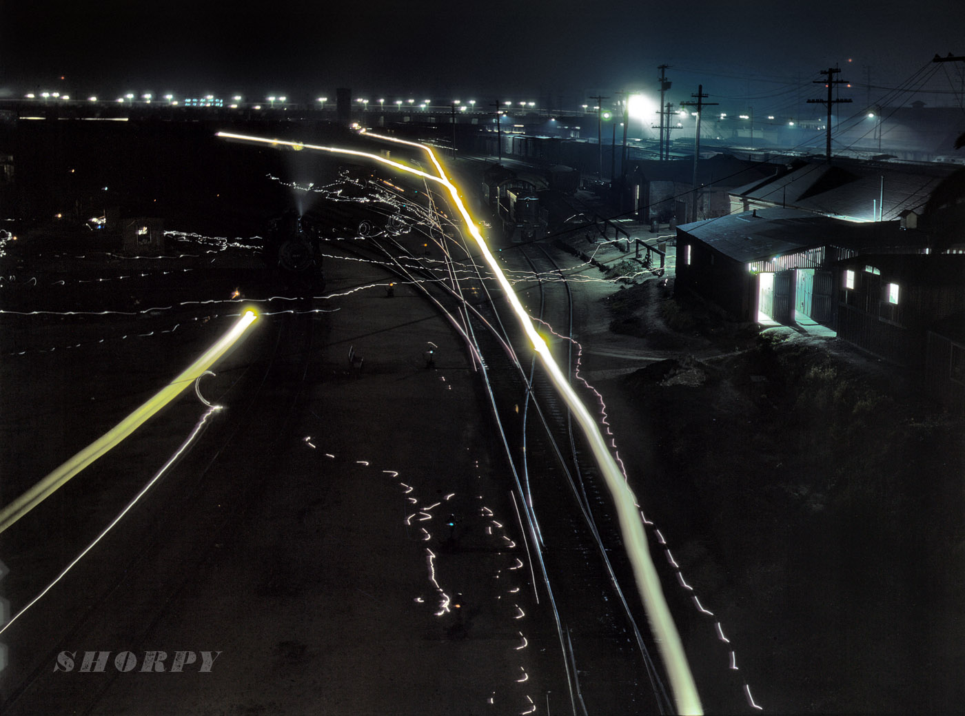 March 1943. Another night shot by Jack Delano, taken on his Santa Fe rail trip west from Chicago in 1943. His description of this scene: "Activity in the Santa Fe R.R. yard, Los Angeles. Due to blackout regulations, floodlights, switch lights, locomotive headlights and lights on the bridge in background have been shaded to cast light downward. Broad streaks of light are caused by paths of locomotive headlights, thin wavy lines by lamps of switchmen working in the yard." 4x5 Kodachrome transparency by Jack Delano. View full size.