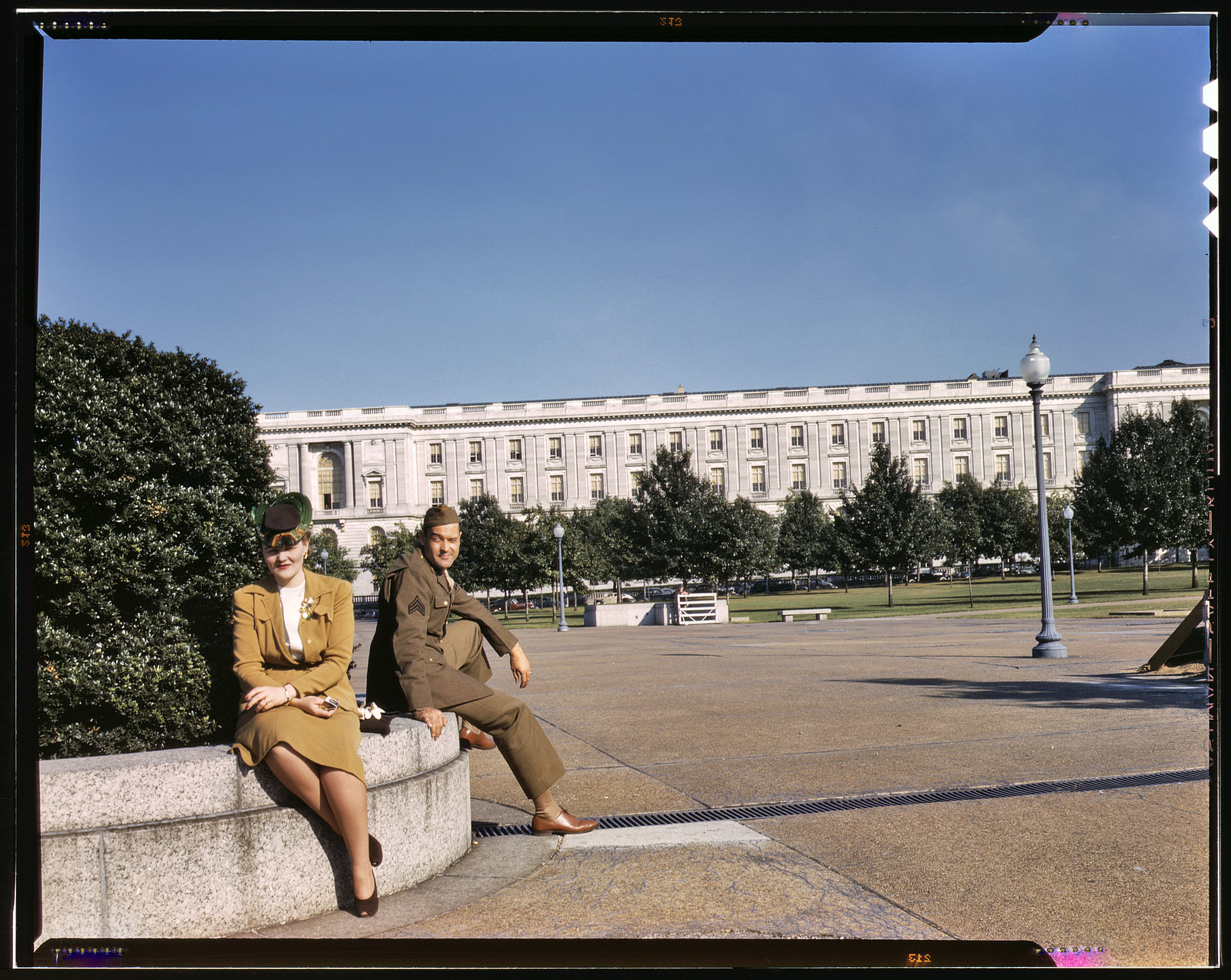 1943. On maneuvers in wartime Washington. "A soldier and a woman in a park, with the Old [Russell] Senate Office Building behind them." View full size. 4x5 Kodachrome transparency, photographer unknown. Office of War Information.