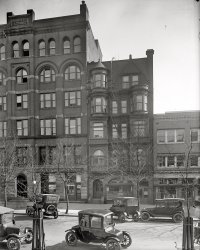 Washington, D.C., circa 1918. "Emergency Fleet Corporation, building exterior." Harris &amp; Ewing Collection glass negative. View full size.
Electric, Gas, and Hay - Oh My!What an interesting time period when it came to transportation. Besides the gas powered automobiles parked on the street, and the Milburn Electric in the center of the photo, you can see the very back end of a horse-drawn wagon in the lower right of the photo. If you look at the plate glass window of the "Builders and Manufacturers" office, you can see the reflection of the horse hitched to the wagon.
FranklinLooks like a Franklin Electric in the foreground, a car about 90 years ahead of its time. Would love to take a drive in one of those today!
On second thought, the carOn second thought, the car might be a Detroit Electric instead of a Franklin,
anyway either would be a blast to drive, although range was limited to 20 miles plus or minus.
3 in a coupeThe 3-passenger coupe was a popular style in that first decade of autos.  The driver had a bucket seat, and the left rear passenger was crammed in behind the driver.  The right rear passenger had more legroom.  Some models included a fold-down jump seat facing backwards in the right front position; this passenger's back was against the dashboard.  
Horse BlanketCan anyone explain the function of the cover over the engine hood on the car in the lower left corner? I'd guess it's there to prevent freezing in cold weather, but it completely covers the radiator face and side vents as well, and I'd think it would tend to starve the carburetor.
1918 Milburn electric carThe car in the foreground appears to be a 1918 Milburn electric car.
ElectricThat's a charming little electric runabout. It's hard to tell whether it's coming or going. 
Electric Car?Is that a Milburn Electric in the center foreground?
This Milburn Electric Model 27 looks similar.
Builders&#039; Exchange BuildingAccording to the Washington Post, this was built in 1890 at 719-721 Thirteenth Street, NW., between G and H (Post, March 20, 1890 p.4; July 25, 1909 p. R2)
Parallel parking?I'm surprised by the haphazard way the cars are parked on both sides of the street. And the jaunty little car center front is a puzzlement. I think I can identify the front by the headlights, but it looks like the passengers are seated facing each other. Where is the steering wheel?
[It has a tiller. - Dave]
Horseless CarriageCan anyone ID the vehicle in the center of the photo? It's intriguing because it looks as if there are two people inside of it that are facing each other.
SteamerI like old cars but I'm not familiar with cars as old as the one in the foreground.  That being said, I'm pretty sure that is a steam powered car
I Tawt I Thaw A Puddy Cat!It appears that there are 3 occupants in the electric car: A woman in an elegant hat, a gentleman in a topcoat, and the chauffeur - a Bedouin preparing for a sandstorm. The last effect may just be due to the tree reflection across the windshield, but it definitely looks like the driver is wearing a mask/muffler of some sort - maybe because to the flu epidemic? Dave, is it time to bring out the ShorpyVision? But the other passenger is missing, because they just dropped Tweety Bird off at the vets.
Razed for ParkingWe tend to think of parking shortage as a contemporary issue - but finding space in the urban environment to park cars has been a challenge since the mass production of automobiles began.  It's hard to believe that a six-story building would be razed to make way for a surface parking lot, but that is what happened.  The Service Parking Corporation razed at least a dozen buildings in downtown to replace them with surface parking.  Other buildings lost to the Service Parking Corporation include the Edward Apartment Building (15th St), Gramercy Apartments (825 Vermont Ave), Marini's Hall (914 E st), and the former residence of President Buchanan (916 E st).



Washington Post, Jan 7, 1933 


Builders Exchange Building Will Pass
Site on Thirteenth Street is Leased to
 Auto Service Parking Company.

Disappearance of the former Building Exchange Building, another landmark of the downtown business section, at 719-721 Thirteenth street northwest, is forecast following completion of an important parking lease for the building's site yesterday by the offices of Carl G. Rosinski and Harvey A. Jacob.
The site of the building, containing 10,093 square feet, has been leased for five years for the Stilson Hutchins estate to Service Parking Ground, Inc, and organization with out-of-town headquarters, which has been in business for seventeen years and is operating more than 50 parking lots throughout the country.

HandlebarsThere is also a bicycle leaning against the tree in the lower right corner.
Motor MuffsThe Fords both have hood covers. Keeping in mind that the engine bay is open underneath, plus the Model T had a low-mounted updraft carburetor, there is no danger of stifling the motor even if the covers were left on during use.
Back seat driverThe electric car was driven from the back seat. If you look, you can see two tillers sticking up on the far side of the car. When the driver gets in, they pull these down across their lap and control the car with them. 
MilburnThe car in the picture is a 1915 Milburn model 15 (their introductory model). Until 1916 most enclosed cars were electric. The electric Coupés usually had a three place bench seat across the back and one or two bucket or folding jump seats that faced aft, or rotated to face either direction.  
(The Gallery, Cars, Trucks, Buses, D.C., Harris + Ewing)