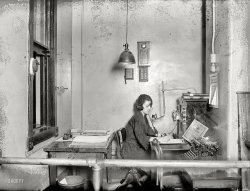 November 1922. Washington, D.C. "Woman's Bureau, Metropolitan Police Dep't. Telephone calls bring prompt attention." National Photo Co. View full size.
Cold OfficeI just noticed that she is on the exterior side of that double hung window.  That really makes this office uninviting!
Behind BarCan't decide if that bar is to keep her in or others out.  In either case, it appears one would have to crawl under it.  At least she has the keys.
Hey! Fish!If this was NY's 12th Precinct, I would expect Wojo and Fish were out on a call. Obviously they modeled the set of "Barney Miller" on this.
ErgonomicsThat particular arrangement is my personal idea of hell.
So spatious and invitingNo expense was spared to accommodate the WB.
[It was an extra-spatial kind of spatiousness. - Dave]
Nothin&#039; like a hairnetTo take away any semblance of sex appeal.  
Call indicator boxI have an oak call box in my kitchen the same as the one to the right of the light fixture; it was once used to summon the servants to different rooms by pushing doorbell buttons. The DC police must have used this one as an intercom of some kind.
Washington &quot;And Nearby Places&quot;What a quaint expression, that!
Not a negative commentDingy, and a lot of it doesn't seem the fault of an old negative.
Cell, PhoneIf this is typical of an office in the DC Police Department, I'd hate to see what the cells in the DC Jail looked like.
 One Ringy Dingy, Two Ringy DingyIs this the party to whom I am speaking?
911 What&#039;s your emergency?We'll have a car out there sometime this week.
Hello CentralGive me Dr. Jazz.
Ruth Buzzi the elderly Lily Tomlin?The large purse is absent!
Giant fingerprint faux finishMaybe Martha Stewart will have a special on how to achieve that in your own police station.
Also, funny how this photo makes even the pencil sharpener look old-fashioned, even though hand-cranked ones are still fairly common.
Everything within easy reach... except the pencil sharpener! That chair will swivel so she can easily use the books on the other table, and the typewriter is well out of the way of the writing surfaces. I've worked in worse.
Security Fire AlarmI love the little iron hammer on the short chain.  Break the glass to get to the fire alarm button.  If a prankster sounds the alarm, just follow the blood trail.  If the fire is real, well, decisions, decisions.
Nearby PlacesGreetings from Bethesda, Maryland, one of those "nearby places." Which unfortunately can now take an hour or more to drive to during rush hour from downtown D.C.  
Guess it's not as nearby as it used to be!
The dark side of the BureauMs. Mina Van Winkle, director of the D.C. Police Women's Bureau, provided this explanation to an audience in Boston in 1920:  The Bureau was organized to enforce "the District's war-time legislation," but "proved so valuable as an emergency measure that it has been made permanent." In 1928 Ms. Van Winkle told a reporter that "Washington is the mecca for all psychopathic women of the nation."
The feature story explained that one of the Bureau's functions was protecting lawmakers "from psychopathic women who flock to the city while Congress is in session with wild and utterly unfounded tales of wrongs done them by prominent men. ... Due to the vigilance of the policewomen, the government officials and other well-known Washingtonians accused of serious misdemeanors often do not even know they have been involved," because the Bureau's policewomen intercept such women, sending some to "some insane asylum" and others home to their husbands, fathers, or brothers.
Depressing dimensionsWhen your office is taller than it is wide, that's not good.
Fish on bun, Jello and milkShe must not have been paid much. From the looks of that sassy hairnet, she had to moonlight as a cafeteria lady.
What, no spittoon?Not fair.
Immaculate PerceptionOf course, this young lady's hairnet was quite common in those days. The cleansers and hair treatments of the day were unsophisticated, which made hairstyling a challenge. Mass production made the fine mesh solution to runaway or frizzy hair available to all women, at a cost most could afford. The hairnets were sold at accessory stores in individual boxes and put out on display, along with the fine gloves and stockings. A great many women, from Bonnie Parker to Eleanor Roosevelt, wore hairnets when they were considered a neat, clean, and feminine beauty product.
(The Gallery, D.C., Natl Photo, The Office)
