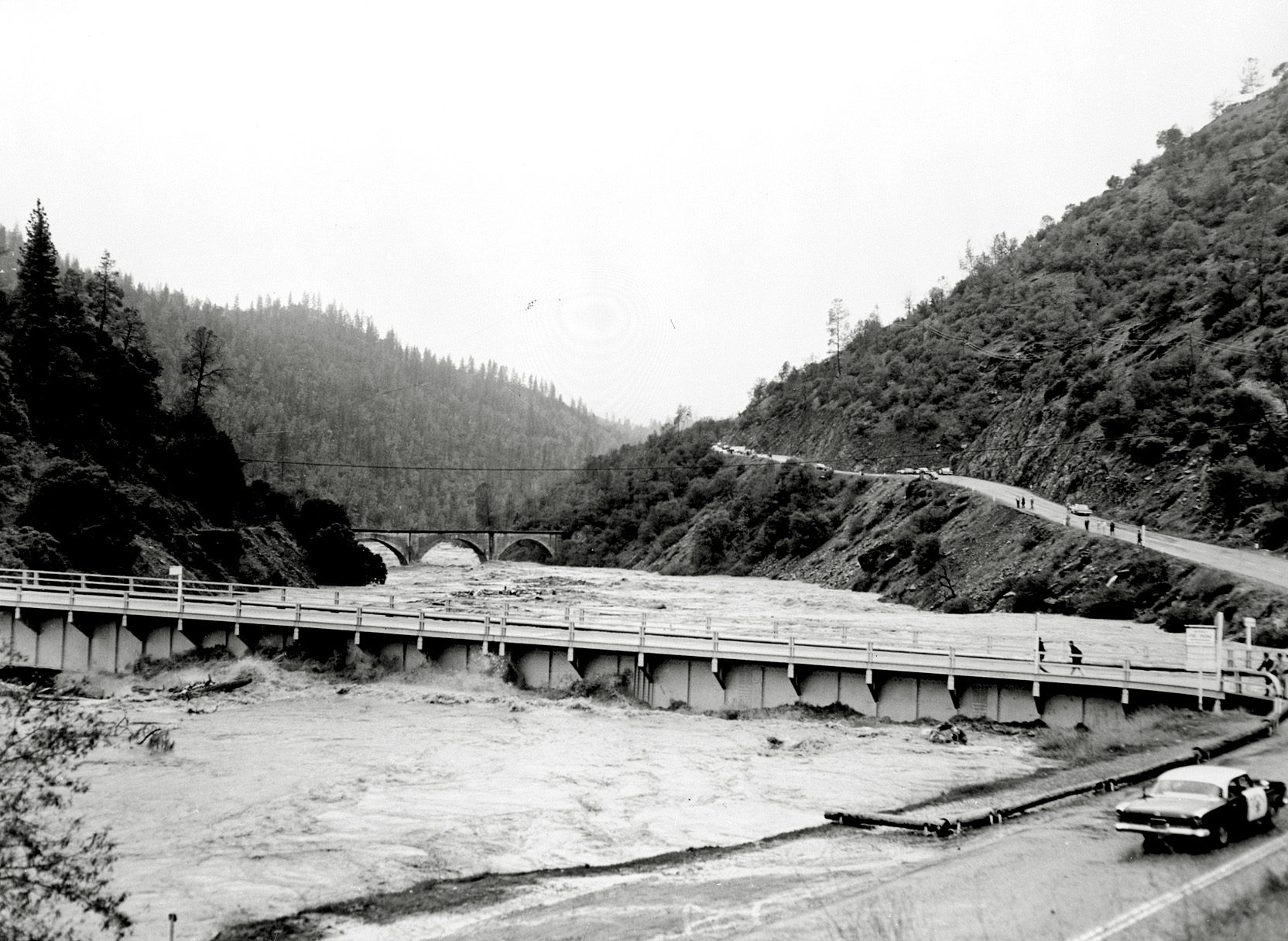 Record rainfall in the Sierra Nevada mountains in December 1964 caused water to breach the yet uncompleted Hell Hole Reservoir Dam located 40 miles east of the City of Auburn, Placer County, California. As the water surge arrived at the Highway 49 Bridge between Auburn and Cool, the Highway 49 bridge was completely destroyed, while the older concrete railroad crossing in the background survives to this day. Norm Sayler Collection, Donner Summit Historical Society. View full size.