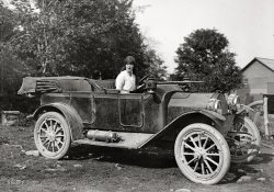 "Woman in automobile circa 1915." From of a series of pictures showing National Photo owner Herbert E. French and friends on motor excursions in the D.C. area. National Photo Company Collection glass negative. View full size.
Gas lamps?That looks like an acetylene bottle on the running board. If that's true, then the headlights have been converted from using calcium carbide to produce acetylene, to being fed from the bottle.
Interesting. I wonder why. Longer running time? More consistent flame/light?
[Convenience and reliability. Prestolite-style pressure tanks began replacing carbide generators around 1906. By the early teens they were standard equipment on many, if not most, cars with acetylene headlamps. - Dave]
A car in its natural environmentLooking at all these fine pictures, I can't help comparing the dirty, mud-covered cars with the pristine specimens in today's museums, and realize that cars back then were not pristine and always clean, but covered in mud and dust. A very interesting contrast. I can hardly imagine what it was like to make a long-distance trip in one.
Road rage?The lady appears to be majorly annoyed about something.  Probably because the men wouldn't stop and ask for directions.
ContrastThere is quite a contrast between this one and Soccer Mom of 1908 from 2 days ago -- setting, cleanliness of the car, demeanour of the driver -- this one looks much more real.
Betty Sue needs a dentistKinda cute and feisty but look at those teeth. I have those lamps and Prestolite tank on my 1917 Buick
Eric
MotoringAnyone embarking on a long-distance trip was well-equipped with tools and expecting to make repairs.  Roughly speaking, one breakdown per trip was the norm.
Just discoveredShe just found out the car has no CD player - what a piece of junk!
Transcript"Don't you dare take my picture right now! We've been on the road all day and I look like heck!"
Good thing she had no way of knowing that people would still be looking at this nearly a century later!
The carPretty sure this is an early (1912-1913) Detroiter Automobile.
(The Gallery, Cars, Trucks, Buses, Natl Photo)