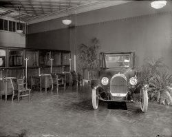 Washington, D.C. "Oldsmobile Sales Co. interior, 1919 or 1920." National Photo Company Collection glass negative, Library of Congress. View full size.
AccessoriesWow! Look at that classic office furniture! Oh, yeah; nice car, too.
Catch PanI guess leaks came with buying a new car in the early 1900s. Can you imagine going into a showroom today and seeing a catch pan underneath it?
FurnitureThings really haven't changed all the much have they? The desks are crammed up real close in the showroom. And instead of PCs on the desks, they have inkwells. They even have the glass offices to run to to get a manager to "approve" a deal.
Drip, drip, dripThis reminds me of the time I let a friend roll his 1975 Norton Commando into my living room to escape a rainstorm.  The first thing I did was slide a cookie sheet under it to catch the oil drips.  Old British bikes had the endearing habit of leaving puddles of oil wherever they were parked.
Your Great-Grandfather&#039;s OldsmobileLooks like a 1920 Model 37A
Floor wax, folks!Must be my hausfrau heart, but it seems like floors in the era needed a good waxing.
OldsI didn't immediately realise how early this is as far as cars go and how quickly they were developing. Just looking at the front of this car...no front brakes, big scrub radius because the kingpins aren't inclined to bring the contact patch into line with them, no carbon black in the tyres etc. I bet that's ordinary plate glass in the windows as well, not tempered. Also, I can't see any front dampers...were they tucked up under the chassis rails or was it simply undamped? Must have been a bouncy thing if so!
Drip LubricationMany vehicles would drip oil as a normal and intended part of their operation.  Metered oil feeds slowly drip oil on various parts that need lubrication.  One very renowned comment aimed at Harley Davidson motorcycles was the misconception that they always leaked oil.  I guess you could call these drippings "leaks".  Intentional ones. Another photo on Shorpy shows a block of oil sight glasses accessible to the driver of an automobile with little thumbscrews on top to adjust the drip rate on each one.
English motorcyclesI had a couple of old Nortons, a Commando and an Atlas, and the comment "Old British bikes had the endearing habit of leaving puddles of oil" reminds me of the old joke:
Q:  What does it mean if your English motorcycle isn't leaking oil?
A:  It's out of oil.
Hey...English motorcycles and cars don't really leak oil....they just 'mark their territory'!
There is the start...The customer's chairs...the salesmen's desks...and the dreaded windows "I'll just pass this by the Sales Manager...you never know"
Anyone wanna bet that is a mirror?I have an Olds of that age and it doesn't leak much oil. Olds had one of the first slanted windshields to prevent glare from headlights from behind. If you think the office furniture is nice, you should see the interior of that car! Pure class! I say that is a mirror to show off the undercarriage.
[Below: A decidedly non-reflective drip pan. - Dave]

(The Gallery, Cars, Trucks, Buses, D.C., Natl Photo)