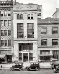 Washington, D.C., circa 1920. "Merchants Bank, G Street N.W." What looks at first like a fairly desolate street scene turns out to have a number of players. National Photo Company Collection glass negative. View full size.
Philatelic scrutinyThe posture of the customer (he's wearing a hat, so I'm presuming) in Harry B. Mason's window leads me to believe he's scrutinizing a stamp album rather than an insurance policy. Then again, perhaps he just spotted the fine print.
Cleveland in WashingtonCleveland motorbike with an old-school kickstand. Awesome!
American Multi-what?From http://thegreatgeekmanual.com/blog/this-day-in-geek-history-december-12 - referencing December 12,  1903:
"The American Multigraph Sales Company of Cleveland, Ohio begins manufacturing the Multigraph duplicating machine, the first commercially successful device to simplify the printing process. It was patented on March 10, 1903 by inventor, Harry C. Gammeter, a typewriter salesman. Consisting of a metal drum with vertical channels running across it, it allows laymen to arrange moveable type with a retaining foot into the channels to roll out professionally lettered solicitation letters."
Horsepower BlanketsTwo more to add to the assortment of engine-warmers seen here on Shorpy.
American Multigraph Sales CompanyI couldn't figure out what a multigraph was, so I looked it up:
From http://stampedout.net/information-022-am_his.html
"1902 - American Multigraph Sales Co. introduces Gammeter Multigraphs.  These were used to mass produce form letters."
Strange at bestOkay, I see the three legged ghost in front of the milliner and the secretary on the second floor.  But what really catches my eye are a few other things.  Like the coffin lining drapes in the bank windows -- sort of a portent of the future?  And the extremely narrow front tire on the truck for rent.  Last but not least, the window dresser of the tailor was ahead of their time.  In the twenties things seemed so ornate compared to the stark plainness of today but this one was "dead" on.  I've seen photos labeled "creepy" here before, but this one gets my vote.
The Merchants Bank

Washington Post, Apr 23, 1918 


Bank to Purchase 5-Story Building

The Merchants Bank, it is understood, has virtually closed a deal for the purchase of the five-story building at 1413 G street, immediately adjoining the building at 1415 G street, which it recently acquired and which it was planned to remodel to meet the needs of a banking house.
President P.A. Drury, it is reported, on good authority, has decided to locate his bank in the building at 1413 instead of 1415 as originally planned, and will remodel this building instead. The bank will retain possession of both buildings, and will lease the first floor of 1415 for stores, which will open on the arcade which passes between the two properties.  The rooms of the upper stories of the two buildings already are connected.  The bank will use as much of the upper stories as may be required to meet its needs, and will lease the rest as offices.
Name That CarOK guys, the first car on our left is definitely a Ford Model T (probably a '17 or '18 touring), the truck is also a T (or a TT?), but what is the middle one?
I always find it fascinating to see what these cars really looked like in real time instead of restored cars or survivors.
The Middle Car is...It seems to be a 1919 Dodge.
"1919 DODGE: From 1916 to 1923, Dodge was built on a 114-inch wheelbase. Until 1919, little change in appearance took place. In March of that year, a four­door enclosed sedan was introduced into the Dodge line. "
Thanks to :
http://www.allpar.com/cars/dodge/dodge-cars.html
Mystery CarIt's a Dodge -- circa 1919 or 1920.
That's the standard, albeit skinny, width for the front tires on the 'T Truck at right. The back axle of the truck seems to be a replacement -- solid rubber tires, different suspension, etc.
Something SocietyCan anyone determine what the writing on the third-floor window of the building on the left reads?  It looks like:  Instructive Visitin***** Society.
[INSTRUCTIVE VISITING NURSE SOCIETY. - Dave]
Multigraph had a long historyThe American Multigraph Company was eventually folded into the Addressograph company, which also provided equipment for mass-addressing of envelopes, brochures, etc. The company was long known as Addressograph-Multigraph (AM), and also as Addressograph-Multilith. They bought the Varityper composing device and existed until the advent of desktop publishing as one of the premier manufacturers of typesetting equipment, AM Varityper.
(The Gallery, Cars, Trucks, Buses, D.C., Natl Photo)