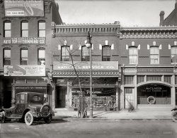 Washington, D.C., circa 1920. "National Service Co. front, 1610 14th Street N.W." Home of 24-hour tire service. National Photo Co. glass negative. View full size.
Seal of ApprovalThese days, we rotate our tires every 6000 miles. So much for the 6000 mi guarantee!
Then and NowView Larger Map
Roadside Service: 50 cents

Washington Post, Apr 6, 1919 

24-Hour Tire Service Begins
J.H. Robison Head of Company
Installing Apparatus for Heavy Work

Twenty-four-hour tire service is something new to Washington.  It has just been introduced by the National Service Company, which has opened a tire store at 1610 Fourteenth street northwest for both solid and pneumatic tires.
J.H. Robison is active head of the new company, which is specializing on the Firestone line of tires and tubes.  Mr. Robison has been for a number of years connected with the American University in the capacity of purchasing agent.  Previous to this he filled a like position for the bureau of mines in the Pittsburgh district.
A 150-ton hydraulic press has been installed for handling the sold tire work.  There is also set up an oxacetylene welding outfit for cutting off old tire bands and tire bases from wheels fitted with the pressed-on type of solid tires.  A service wagon, specially designed, with an overhead trolley and chain hoist for handling heavy wheels, is in operation.
For the pneumatic tire business a complete vulcanizing plant is being installed and a service wagon that will answer telephone calls from anywhere in the vicinity of Washington, night or day, is on the streets.





1920s Taggers!Apparently, graffiti was a problem back in 1920, as well. Take a close look at the lamppost. Danged whippersnappers!

Free AirProprietor: "That'll be fifty cents for the tire fill."
Customer:   "I thought the air was free."
Proprietor: "It is but there's a non-waivable atmosphere-to-tire transfer fee."
P.S. The lady in the window is reverse trick of the eye - she's real but looks like a cardboard picture.
[She is covered with mold (on the emulsion, at least), which gives her that washed-out look. - Dave]
Some things never changeI have a fondness for company logos that have remained in use for decades.  The Firestone logotype hasn't changed in at least 90 years.  That's a good one.
Auto BedsI guess Motel 6 leaving the light on for you wasn't around yet!
Dapper CasperThe ghost in front of 1612 appears to have had his shoes shined.
ChillinI like the milk bottle on the window ledge -- cheaper than buying an icebox, I guess.
(The Gallery, Cars, Trucks, Buses, D.C., Natl Photo, Stores & Markets)
