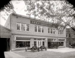 Washington circa 1920. "Steuart's Garage, 12th Street N.E., exterior." Last week we saw the interior. National Photo Company glass negative. View full size.
LampsThose are the cowl lamps sitting on top of the frame. The base model Fords of that era had kerosene cowl and taillamps, nondemountable rims and no electrical system. They were crank start only, no battery. The headlights ran directly off of the magneto. Dim at idle and prone to burn out at high (for a T) speeds.
Reversal of printHey Dave, any idea why the printing on the office door glass is reversed?
[We are looking through a window at the back of one of two central (open) double glass doors. - Dave]
Store Across the Street...Looks like the reflection in the large plate glass window shows that Leonard P. Steuart (Got the name from the earlier interior photo) had a similar building across the street. This reflection is probably the 141 address shown on the parked truck.
The address on the parked truck advertises 141 12th Street which would make sense if the even numbered 142 Google map image address shown is correct.
The 141 address and "151 Office" reflection would then be on the camera side of the street.
I am still having a bit of trouble visualizing the "151 Office" reflection though. The camera must have been located farther back into the store across the street and was shooting through a closed glass door with the number on it.
["151 Office" is not a reflection. See note below. - Dave]
The name is still there...View Larger Map
Gas tank seatWe've come a long way in auto safety. Imagine sitting over the gas tank. 
Ice cream parlorWhat a pretty building. It looks more like an ice cream parlor than a garage!
I don't understand why the lettering on the office door is backward. Anyone?
Running on emptyWhat a nice-looking shop! I'll bet these simple Model Ts were a cinch to work on compared to the overcrowded engine compartments of today.
As for that gas tank; I had an old friend that owned a "T" in the 1930s. He said that when the fuel got low, the engine would customarily quit when going up a hill. He would then roll the car back down the hill, turn it around and drive backwards up the hill because the gas tank pickup tube was towards the front of the tank. Backing up the hill would allow him to scrounge the last bit of fuel to make it home.
151 - OfficeVery cool picture.
I'm curious why the "151" and "Office" that appear to be on the door between the two cars is reversed ...
Cowl-chassis trucksA couple of cowl-chassis trucks are parked out front, waiting for commercial bodies to be fitted.  It looks like the factory-supplied taillights are temporarily attached to the top of the frame rail, similar to what Ford still does today, on their Super Duty cab-chassis trucks.  Also, the building across the street, reflected in the front windows is still there (swing the Google Street View around, to see the telltale five windows above the front doors).
BYOB: Bring Your Own BodyInteresting insight into the evolution and birth of the coach-built era of automotive history, where you bought the running gear from a supplier of your choice, and sent it to a coach-builder, who created a body for it, based on your specifications.
Cars now worth millions (such as Deusenbergs) were often built that way. But you don't think of the lowly Model T in the same thought bubble as a Talbot-Lago. 
Some kinds of cars were still created that way such as ambulances out of station wagon running gear, until recently. And trucks are still sold as cab and rails with a second supplier adding the box or crane or whatever it gets. But short of an aberrant Brabus, this kind of auto-making seems to be a lost art. Suspect those two T's were getting wooden bodies after they sold.
Wonder if you could buy a current Ford car (not truck) minus the body, and coach-build it? Bet the folks who do crash tests nixed that option for people today.
(The Gallery, Cars, Trucks, Buses, D.C., Natl Photo)