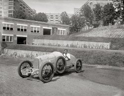 Washington, D.C., circa 1920. "Donnie Moore in Duesenberg." Last seen here a year ago. National Photo Company Collection glass negative. View full size.
Location, Location?I'd guess a large apartment complex in the NW side of D.C. The units on the end of the wings have sleeping porches rather then balconies.  We're seeing it from the back so hard to pin down exact location!
[It's the Wardman Park Hotel on Connecticut Avenue, built in 1918. With 1,000 rooms, Washington's biggest. Later the Park-Sheraton. Torn down in 1979 and replaced by the even bigger Marriott Wardman Park.- Dave]
No StoppingThis looks like a heavy, powerful car. There are no brakes in front.  Modern cars have 70 percent of their braking power in front. Perhaps this car was only intended for the wooden racetrack shown earier this week.
Give it up for Donnie.I'd give my left wheel nut for that car.
Drive Slow!Is that a dare?
Friction shocksIt has friction shock absorbers front and rear.  Note the discs with the stars at each corner for adjusting the preload.
DRIVE SLOWNice contradiction.  Body work on the car appears less than concours.
Keeping the wall upI find it amusing how many of these old photos have a guy in them, leaning up against a wall or door frame exactly like the guy in the background here.  
That car must have been one wild ride.  With his low seat position it seems that he'd have poor visibility of the road ahead of him too.  
On the road againThe one thing that jumps out of this picture is that this high performance racecar is wearing D.C. license plates. You could drive it around town -- loud pipes, no headlights, just go for it. Must have been a ball to drive.
That steering wheel is enormous!And how did he ever see over that dash and hood?
No power steering needs big steering wheelThe large wheel provides a lot of mechanical advantage when turning. Reminds me of buses when I was a kid. They all had enormous steering wheels and the drivers would start their turns early and turn and turn and turn the wheel to get around corners. If you ever saw Jackie Gleason in the Honeymooners in the driver's seat of his bus, that is the type of steering wheel buses had way back then.
Duesie x 2A few years back one of my clients, a high roller, attended a classic auto auction and came home with two Duesenbergs, having parted with $1.5 million.
Get him a phonebookHe can barely see over the hood. Is that for better aerodynamics or is he just short?
A minor observation....This car may well have been driven on a boardwalk surface, but if so then it was also equipped for racing on a dirt track: note the mesh grille protecting the radiator from being damaged by flying stones.
More to the Point!I wonder where Donnie's Duesie is now -- somewhere in Laurel near the old race track? 
Incredible stuffNobody was braver - more insane? - than the board track racers. If you want a definitive look at this era, read Dick Wallen's terrific book, "Board Tracks: Gold, Guts and Glory," which is still in print. 
(The Gallery, Cars, Trucks, Buses, D.C., Natl Photo, Sports)