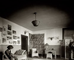 Washington, D.C., 1920. "National Photo Company, front room." View full size. Glass negative: National Photo Company Collection. Styling: Morticia Addams.
BackdropIs that a camera on the far right?  I bet a lot of portraits were taken in front of that wall of sharp implements.
Now that is some seriouslyNow that is some seriously weird-looking furniture. The whole room has a very uncomfortable feel to it IMO - two thumbs down for the interior designer.
Early GuestUpon spotting the odd collection of weapons hanging on the wall the early guest began to wonder who has invited him to dinner. Or if it was him who is the dinner...
My voteIs two thumbs up!!! I just love the oddity of it all and I cannot believe it is the waiting room of a business office!! I don't think I would leave for hours - just looking at all of the photos and weaponry and antlers, OH MY!
My thought...is that the photographer put all the photographic staging pieces in the front room.  I also think it's a good idea to display some curiosities for people to look at whilst waiting.  I wish more businesses would do that today. 
Love The Title!I laughed out loud at the title of this photo! Comfy, indeed! In that room, I imagine that the furniture is made out of human bones! Although bamboo might be closer to the truth. Does anyone want to guess what the subject matter is in the photo next to the antlers? It appears to possibly be a close-up of some sort of animal dung? But, I'm sure I am wrong about that, too. I hope...
Kathleen   
Charming photoMy guess about the photo next to the antlers is that it's a coiled snake.
I get a whole King Kong/Skull Island vibe from the furniture motif; I wonder if Merian C. Cooper had visited the National Photo Company 13 years before? I would definitely make room in my place for that vanity/lamp combo, although I'd be real careful getting up from it.
Mighty Hunter?This is an odd juxtaposition of styles. That desk looks painful. It actually looks more like a vanity table. Maybe this is a young couple with the usual starting out mixture of his manly trophies and her teenage girl bedroom furniture.
[This is the reception area of a business office. And that may well be Herbert E. French himself on the couch. - Dave]
(The Gallery, D.C., Natl Photo)