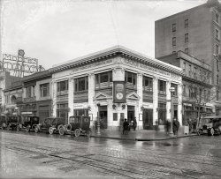 Washington, D.C., circa 1922. "Security Savings &amp; Commercial Bank, 9th and G." National Photo Company Collection glass negative. View full size.
All that&#039;s left today...is the storm drain inlet in the curb.
Christmas ClubMy mom always urged us to join the local bank's Christmas Club as very young kids because even though we received no formal allowance as kids, we could somehow manage to scrape together 50 cents a week and in early December, we would receive our check for $25.00.   How wealthy and prosperous we would feel when we received in the mail that official bank check with which to buy gifts for our parents, siblings and best friend.  When I think back to how kind the bank tellers were (never annoyed) when we would come in with our coinage and bankbook and how they would take the time to enter our deposit, rubber stamp the page and assure us of how smart we were, we received personal service and attention with no extra charge.  With 3% interest on our money that was more than the savings accounts give us today.    Ya' really have to wonder where it all went wrong and we got to where we are today.  No, I wouldn't want to go backwards, but there was a time when every person mattered, and still the banks made sufficient profits.
FascinatingI like the fact the two clocks, one on what I presume is the Rialto, are very close in time, and someone who appears to be a Louis Abraham was next door to the bank buying gold and diamonds. Finally, I notice the bank was paying 3 percent on Christmas Club. Of course in recent years, banks just simply couldn't do that.
Parallel ParkingThese drivers could really park in tight spaces!
PopupYou can see a man's head on the right near the sidewalk. According to my learned co-worker, he is utilizing a freight elevator.

Hanging out..I love the wise guy hanging out, leaning against the tree without a care in the world!
Street Car TracksNotice the streetcar tracks with the slot in the center for the 3rd rail plow for power pick up.
Overhead trolley wires were not allowed in DC
Head above the parapet.Did anyone notice the guy emerging from the cellar to watch the ghosts pass by?
Seven years laterthe same corner was probably crowded again, but for substantially different reasons.
Cable Car?I think D.C. had cable cars, not electric trolleys.  That line in the middle would be for the car's cable-gripping apparatus, not an electrical contact.
[These tracks are for electric streetcars.  D.C.'s cable cars were replaced by an electric system in the 1890s. - Dave]
Christmas Savings, did I make a mistake?I have to pay one year = 52 weeks every week 50 cent and I get $25 only? Where is my profit?
[50 weeks, not 52. - Dave]
What is the odd dark globeWhat is the odd dark globe fancy street light in front of the bank?  Is this a fire or police call box?
[Police call box. - Dave]
(The Gallery, D.C., Stores & Markets)