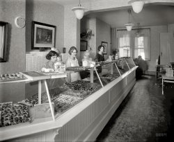 Washington, D.C., circa 1922. "Benj. Franklin candy store, 13th Street N.W., interior." National Photo Company Collection glass negative. View full size.
QuestionWhy do visions of candy on conveyor belts and Lucille Ball go through my head while looking at this picture?
While You Were Away 

1922 Advertisement


"Benj." Franklin Candies
Pays Us a Visit and Sample our Famous
BUTTER CREAMS and GLORIA ROLL

Why is everybody talking about "Benj." Franklin Candies?  Because we believe our candy is the finest and purest in the city of Washington; our candy is all home-made &mdash; made the same as Mother would make it in the kitchen at home.  A trial will convince you of its superiority.  Remember the Address &mdash; 517 Thirteenth Street  &mdash;  Opposite Palace Theater.
Branches: Baltimore, Richmond, Dayton, Fairmont, Morgantown, Wheeling.



Update: Don't wait! It won't be here long!


1923 Advertisement





Have a gloria roll and make  yourself at homeIt looks like they've set up shop in somebody's living room.  There's even a coat hanging over the doorknob. 
The &quot;clock&quot;Cover plate for a stovepipe hole.
Those &quot;Candy Girls&quot;look shell-shocked.  Was is something in the candy?  The working hours?  The fact that they were forced to pose forever while the photographer did his thing?
What I want is To be locked up in this place overnight with a half gallon of cold milk. They would find me the next morning on the floor lying on my back, distended belly with an empty milk container, chocolate all over my face and mumbling incoherently.
All in the FamilyThe two center women have the same nose and head shape.
I think this is a mother, father (in the back) and their two daughters.
I also think this is not what those two sisters wanted to be doing on their summer vacation. That accounts for their "are you done yet" teen-aged glares.
Did the &quot;Heat&quot; do them in?Is that gent by the window on a telephone?  Old time candy stores often served as a front for a bookie operation. Those oscillating fans couldn't protect the goods from the brutal heat of a D.C. summer.  Three thousand pounds of leftover coconut?  My Mother worked in the Sander's candy factory in Detroit, Michigan for a spell in late 1930's.  She was told she could eat all the candy she wanted as she worked.  After the first day she had her fill.     
They&#039;re really...Zombies!
Ma? Meh.No wonder this store went bankrupt.  If they advertised candy as "the same that Mother would make" at home, then I'm taking my sweet tooth to a place where they actually know how to make candy.
(The Gallery, D.C., Natl Photo, Stores & Markets)