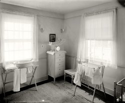 Circa 1920. "Nursery." National Photo Company glass negative. View full size.
NurseryI can smell the baby powder.
HazardsI count 6 or 8 strangulation hazards for toddlers. Babyhood was more dangerous in those days. 
Strictly germ-proofthis bare white interior reminds me of the old poem:
Strictly Germ-proof
THE Antiseptic Baby and the Prophylactic Pup
Were playing in the garden when the Bunny gamboled up;
They looked upon the Creature with a loathing undisguised;—
It wasn't Disinfected and it wasn't Sterilized.	 
They said it was a Microbe and a Hotbed of Disease;
They steamed it in a vapor of a thousand-odd degrees;
They froze it in a freezer that was cold as Banished Hope
And washed it in permanganate with carbolated soap.	 
In sulphurated hydrogen they steeped its wiggly ears;
They trimmed its frisky whiskers with a pair of hard-boiled shears;
They donned their rubber mittens and they took it by the hand
And elected it a member of the Fumigated Band.	 
There's not a Micrococcus in the garden where they play;
They bathe in pure iodoform a dozen times a day;
And each imbibes his rations from a Hygienic Cup--
The Bunny and the Baby and the Prophylactic Pup.
Scrupulously CleanSo clean and tidy. Love the oval white enamel baby tub, although the rickety canvas-top stand wouldn't pass OSHA standards today! Quaint wicker baby scale on top of little chest. And it looks like Mother has washed out some gowns and receiving blankets, drying on the clothes rack. Room itself is so placid and restful; undoubtedly the Baby was loved &amp; wanted.
Hold still, kidThe sleek five-drawer dresser would look good in a modern home.  The flimsy scale on top of it, though, is a disaster waiting to happen.  One good wiggle and the baby would slip right off.
Coming in or going out...It has been said we enter the world toothless, bald and crawling around in diapers and we pretty much leave the same way.  Except for the scale, this is similar to single rooms in a nursing home.  Sorry to be a wet blanket, I usually look at the sunny side of life, but this is a serene and sunny room and would be a happy place for a new baby.  So maybe we all go back to our happy place, huh?
Tinctura opii camphorataRE: "Hold still, kid"
That's what paregoric was for!
Look how convenient the electrical receptacle is, no bending over. And one less hazard for a crawling baby.
Cloth diapers!I love the cloth diapers on the drying rack.  I hope the mother had an electric washer with a wringer on it, to wash the diapers.  My grandmother had to live with her in-laws when my mother was born. Her mother-in-law had a wringer washer, but wouldn't let Grandma use it, so she had to take the diapers down to the "crick" and wash them by hand.
When I was young, I took care of a little old lady who was born in 1883, and had twelve children.  One day, I commented that she must have spent many, many hours washing diapers by hand.  She got a smile on her face and said, "But it was an easy washing. Besides, the hardest thing was getting the money to buy the cloth to make the diapers." I thought of that often, years later, as I washed the store bought, pre-fold diapers for my babies, with an electric washer and dryer, and never felt like it was a burden.
(The Gallery, Natl Photo)