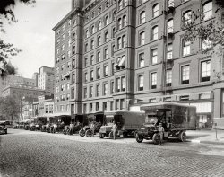 Washington, D.C., 1923. "W.B. Moses &amp; Sons, F and 11th Sts." Note the unusual circular windows at the top of the building. National Photo Co. View full size.
Harrington HotelWashington's oldest continuously operating hotel. 436 11th Street N.W. (corner of 11th &amp; E) Opening day: March 1, 1914
http://www.hotel-harrington.com/index.htm
Drivers, Take Your MarksLooks like an old gran prix style race start. Those round windows were the first things I noticed. I believe there was a similar round swiveling window in the attic of the Griswald home in Christmas Vacation. Just a fact taking up space in my brain.
Also notice that long straight-up fire escape. Seems like the stairs type would be safer in a panic situation. There are also flag holders outside many of the windows.
&quot;Fire Escapes&quot;Those are probably for getting into the building, not out. The ladders are fire-control access to the various floors. There are standpipes on either side with a connection point at each floor and two supply connections at the bottom.
AwningsWho did you have to be to get awnings on your windows?
[A shady character? - Dave]
Top heavyThose trucks look so top heavy. I bet a lot of them ended up on their sides during this era.
W.B. Moses &amp; Sons Washington Post, Jan 4, 1914


 W.B. Moses &amp; Sons
 The History of a Great Washington
Business Establishment

Although Washington makes no boasts of surpassing the great commercial centers, it should be a matter of no little satisfaction and pride to Washingtonians to know that here in the Capital is located the largest exclusively retail furniture, carpet, and drapery house in America - the widely known establishment of W.B. Moses &amp; Sons.
During the civil war, over fifty years ago, Mr. W.B. Moses, who had for a number of years been engaged in the furniture business in Philadelphia, came to Washington upon the dissolution of the partnership in which he was interested in the Quaker City, and rented a small store on D street, between Sixth and Seventh streets.  He had received, as his share of the stock upon dissolution of the Philadelphia firm, about three carloads of furniture, and with this he opened business here.  He met with such demand for goods that most of the stock was sold on the pavement before he had time to move it into the store room.
Within a few months he rented the three upper floors of the building at No. 508 Seventh street, and, several months later, he rented the large building at the corner of Seventh and D streets, which he occupied in addition to the other establishment.  Meeting with continued success, and his business constantly increasing in great strides, in 1869 he secured the old hotel at the corner of Seventh street and Market space (on the site of the present Saks building) know as the "Avenue House."  Here Mr. Moses executed a business innovation which attracted attention over the United States.  He fitted up the parlors, libraries, dining rooms, and bed rooms completely, decorating and furnishing them in every detail and particular, and in many different colors and combination of colors, in order that almost any one's taste could be satisfied. As a result of this venture, it was no unusual occasion when a patron would say, "Duplicate this apartment" or "that apartment" - meaning the furnishing of an entire house like the sample shown.
His business continued to improve until, in 1884, he purchased the property at the corner of F and Eleventh streets, and began the construction of the present building, now occupied by W.B. Moses &amp; Sons. This building was started in June, and was open for business in the following October.  The architect was Mr. A.B. Mullett, who designed the State, War and Navy building, and Mr. John Howlett was the builder.  They built well, and it was said they completed the building in less time than any building of like size had been constructed up to that time.  Mr. Moses conceived the practical idea of going two stories underground; and as this was a radical departure for Washington, the citizens, when the observed the great excavation being made, predicted bankruptcy and complete failure for the enterprise, particularly as there was no business to speak of in that day on F street.  W.B. Moses &amp; Sons were the pioneers.
Shortly after, through the efforts of Mr. Moses, Woodward &amp; Lothrop removed from Pennsylvania avenue to the present location at the corner of Eleventh and F streets, and property on this short business street has been greatly in demand.  In 1884, $2.50 per square foot was the prevailing price; a short time ago a lot 30 by 75 feet, near the Moses building, could not be bought at $90 per square foot.
The original Moses building was seven stories high, with two stories underground making nine stories, and covered a space 50x100 feet.  In 1887 an additional piece of property on F street, size 25x100, was secured, and built up seven stories to match the main building.  Still another addition was put up on the Eleventh street side 50x100, in 1889, matching the original building.  Again and 1898, still another building was put up on the Eleventh street side, 45 feet front; this addition was built up ten stories, and is absolutely fireproof.  This later addition to the Moses establishment was designed to accommodate the shops and manufacturing departments of the store.
The present establishment of W.B. Moses &amp; Sons is one of the handsomest and most complete stores of its kind in the United States, and the largest house devoted to the sale of retail furniture, carpets and draperies.
In their factory, which is by far the largest in Washington, there is employed a great force of artisans and skilled workers, cabinet makers, upholsterers, &amp;c. The firm furnishes estimates on furnishings, draperies and decoration, and where desired decorative effects in the color will be designed for the approval of patrons.  This branch of the work is in charge of a skilled artist.
One of the store events looked forward to by many thousands of Washington families is the "Annual January Sale," an occasion when prices are quoted which are a genuine surprise, and are away below the real values of the merchandise.  This sale is scheduled to begin tomorrow, Monday, January 5th, full details of which will be found in today's papers - Advt.

FurnitureI actually just inherited a piece of furniture with a label on the back saying W.B. Moses and Sons Washington D.C. Causing me to google the name. I am trying to find out approximately how old the piece is. Any Ideas when this store closed its doors?
Moses ClosesW.B. Moses &amp; Son closed up in 1935.  Press accounts in May 1935 indicate the store is still open.  However, on September 29, 1935, an article in the Washington Post reports the former site of W.B. Moses &amp; Son at Eleventh and F has been leased as office space.  A few ads in early 1936 announce the auction of oriental rugs: "Balance of the former stock of W.B. Moses &amp; Son"
Moses MaterialI have found a beautiful piece of cloth in my aunt's belongings, with a store tag pinned to the cloth -- "W.B. Moses &amp; Sons, Washington D.C." It is the most exquisite, and very dense piece of red velvet, with a price tag of seventy-five cents. I believe the cloth belonged to her mother who took this cloth to China and back to the U.S. somewhere around 1920. I can’t imagine the price it would bring today. It would have been nicer that it was a piece of furniture but I am happy to have the cloth. The store must have been grand.
+77Below is the view of 11th Street looking south from F Street taken in April of 2010.  The Hotel Harrington can still be seen in the distance on the corner of E Street.
&quot;Ask The Man Who Owns One&quot; (or Two)The two trucks on the far right are Packards.
The truck with license plate "36 205" is a circa 1913 model and the truck with license plate "36 204" is a circa 1911 model.
The primary identifying feature is the radiator grille which is a widened version of what was used on Packard passenger cars starting in 1905.
Packard trucks were made from 1905 - 1923.  In 1912 a Packard truck carrying 3 tons was driven from New York to San Francisco in 46 days (July 8th to August 24th).  
Many of Packard's large automobile chassis were also used for commercial purposes such as hearses and ambulances.  These commercial auto chassis were available well into the 1950s.  Packard's automobiles were made from 1899 - 1958.
(The Gallery, Cars, Trucks, Buses, D.C., Natl Photo)
