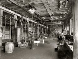 Circa 1916. "Hackett Motor Car Co., Jackson, Michigan." National Photo Company Collection glass negative. View full size. Another view here.
Old iron!Let's see ... Two W.H. &amp; J. Barnes Drilling Machines (or possibly Rockfords, but they don't have back gears) and one of a different model that may be an earlier or smaller version, or a different brand entirely.
The near machine is a Rockford (no relation) No. 3 hand miller, and the small pedestal grinder just past it is, I think, a Buffalo Forge- the work rests are sitting on the crate in front of it.
I think the shaper is a Gould &amp; Eberhard 16", but I'm probably wrong. Can't see enough of either lathe for an ID, but the horizontal mill toward the back right is almost certainly a small Brown &amp; Sharpe.
All that and some gas lamps, too!
Mind your fingersShops like this one are engineering marvels.  The big belts and shafts, the gaslights and machinery all on a safe oil soaked wood floor littered with stock, fascinate me.
Drive beltsIt must have been a job keeping those belts from spinning right of their pulleys. Alignment must have been critical. I imagine that there was a trade devoted to that task. And when a belt snaps and flies off there were whoops and hollers! All right, all right. Get back to work.
Wow!  OSHA would have a stroke!Riveted water heater tank, overhead belts, no guards on grinders, parts all over the floor, overhead gas lighting and, I'm sure, open containers of solvents somewhere in the mix.  Yassir, 'nuf stuff in there to keep the OSHA inspector busy for days!
The Winner OfThe Poor Housekeeping Seal.
Thoroughly Modern PulleyThis appears to be a very modern shop for its time, with the gaslight and the power-driven machines. When I was young we used flat belts like these to power our buzz saw -- we attached it to our John Deere Model A tractor. When those belts broke the result was sometimes tragic.
Alignment not-so-criticalAT wrote: "It must have been a job keeping those belts from spinning right of their pulleys. Alignment must have been critical."
Actually, with flat leather belts, the pulley alignment is not a critical factor in their performance.  Usually the flat pulley has a slight crown on the surface which helps to keep the belt centered on the pulley.  The torque delivered comes strictly from the friction between the flat surface of the belt and the pulley face.  
The more modern v-belt requires precisely aligned pulleys because the friction occurs between the edges of the belt and the walls of the pulley grooves into which the belt is forced by drive tension.
More on Drive BeltsDrive belts were not prone to come off their pulleys.  One of the things we studied in engineering when I went to school was how they worked.  The pulleys have a slight crown on them. As the moving belt starts to move sideways off a pulley, the crown causes it to move back.  We analyzed the mathematics of the process.  The belts are self-centering.
Re: Belts and shafts...Actually, you'd be surprised how easily those line shafts could be set up.
Typically the shaft supports had some method of adjustment- quadrant screws, slots, etc. That mad it as simple as bolting the bracket to the board or joist, run the shaft through it, and tweak the screws 'til it's straight.
You can see the adjusters fairly clearly on the left-hand post-mounted shafting, and on the nearest-right ceiling shaft that powers the pedestal grinder.
The pulleys are almost as easy. Yes, they need to be somewhat closely aligned, but they're slightly crowned and typically wider than the belt itself. That and the distance allow them to be misaligned by quite a bit and still work reliably.
Setting the tension was reasonably easy too- cut the belt to length, press in new clips. pin back together, done. If/when the belt stretches or breaks, just cut and add another clip.
It was actually surprisingly efficient for the time.
Overhead Drive BeltsMy father used to work in the Erie RR maintenance shops as a teenager back before WWII.  He was telling me one day about all the overhead belt drive machinery.  The belts are all leather and apparently making, fitting and adjusting new belts was a skilled trade in those days.  He remembers a company that would come in about once a year to inspect, adjust and, if necessary, replace worn belts.  This was all tied up with getting the drive gear ratios adjusted properly for each piece of equipment, and the drive wheels placed properly.  According to Dad it was not a task you took lightly because an improperly adjusted belt or worse, a broken belt, would have serious consequences.
Under PressureThat's an an air tank, not a water heater. The compressor is to the right.
Dad&#039;s old shopFor many years my dad was a machinist for the NYC Department of Sanitation. I'd visit him at his job in Bed-Stuy. It always had a smell of grease and oil. I loved that smell. 
Back in those days the they would burn the trash right on site in these huge furnaces. I remember the shop always being nice and toasty in the winter and hot as hell in the summer. 
I always thought it was the neatest place to be.
Electricity isn&#039;t far behindMaury Klein has a great book called The Power Makers which describes the race to industrialize using the power of steam (which clearly was the driving force of this shop), the rate at which steam depleted forests and then led to widespread use of anthracite coal, and finally to Edison and Westinghouse's battles to electrify the country.
Highly recommended, a very engaging study of the key decisionmaking and makers behind the US industrial revolution.  His description of the mighty Corliss steam engine will make you weep.
(The Gallery, Cars, Trucks, Buses, Natl Photo)