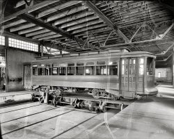 Washington, D.C., circa 1932. "Capital Traction Company trolley in car barn." 8x10 safety negative, National Photo Company Collection. View full size.
I Should Have KnownWhen I saw this Shorpy photo, I became instantly curious as to the origins of the offbeat name, "Capital Traction." A little research revealed that the company and its name were the brainchild of our US Congress.  Now it all makes perfect sense to me.
[There were hundreds of traction companies in the early part of the century, the traction coming from a moving cable under the street. A lot of them eventually adopted electric propulsion or switched to buses. - Dave]
I wonderIf O. Roy Chalk had a framed copy of this picture somewhere in his offices when it was D.C. Transit? Neat picture, thanks, Dave for the posting.
CTC 679Back to the library:
Capital Traction 679 was part of a group built by Jewett in 1911, Nos. 621-700. Succeeding Capital Transit renumbered them 207-286.
These were part of the largest group of DC streetcars built by Jewett from 1910 to 1912. This series of cars was scrapped between 1945 and 1947.
Note this is NOT a "trolley car." There is no trolley on the roof. Many of this series never had a trolley pole; they ran off the conduit inside the slot between the running rails, indicating this car spent its career running in the DC inner-city.
 The conduit system was mandated by Congress to eliminate unsightly wires along DC streets.
[Fascinating research! Generally speaking, a trolley car can be "any wheeled carriage running on a track." Over the years, Capital Traction operated both electric and cable-traction streetcars. Washington's only cable-car loop ran out of Capital traction's car barn in Georgetown. - Dave]
Transfer TableThe car is sitting on a transfer table.  Transfer tables were used to switch cars using a much smaller space than a traditional yard with turnouts.
Going Sideways.I see the Capital Traction car shown on the transfer table collects it's its electric current from a shoe suspended between the rails in the centre of the track.
The rectangular checkered "manholes covers" allow maintenance of the current conductor under the pavement level.
This method eliminates all the above the surface of the street, trolley wires, support cables and poles, presenting a cleaner street view.
(Maybe Dave can find a photo of the current collection shoe hanging beneath a streetcar? I understand there were concrete pits in the streets where the shoes could be applied and removed at locations where streetcars so equipped could change from shoe operation to trolley pole and wire operation in suburbs.)
[See this post. - Dave]
The car shown has only two traction motors on the inner two axles.
The wheels on the outer two axles are of a smaller diameter than the inner wheels with the motors, and the journal boxes on the outer axles reflect this and are closer to the road surface.
Apparently some versions of this style of two-motor truck was more prone to derailment on curves than the standard streetcar truck with all the wheels the same diameter.
Some eight-wheel cars had only two motors for use in flat cities, many more cars had four motors, some cars had no motors at all and were trailers pulled by the motor car coupled ahead, with an electric jumper cable between the cars for the towed trailer's lights and so it's its Conductor could signal the Motorman in the front powered car.
The electric traction motor and it's its gearing to move the transfer table at right angles to the streetcar tracks on each side of it is visible to the right below the wooden walkway.
The controller for the table is to the left, and is similar to one found on a streetcar.
The power for the transfer table is picked up from power rails parallel to the table on the end similar to the "third rail" found in subways.
True Dave, butI'll grant that most would call this a trolley car. And CT did run some cable lines, but the cable was long gone by the c.1932 time of photo.
The slot in DC track was for spaced contacts which were swept by a skate or plow attached to the bottom of the car. The skate was always in touch with at least two contacts. [I believe New York City had the same conduit system.]
Any former residents of DC region about 60 years of age will remember the "plow pits" located near the District line; here the pit man would remove the skate or plow from under an outbound car while the car's crew would raise the trolley pole for the ride into the suburbs. Inbound cars reversed this procedure.
All of this is to make the point that CT 679 is an electric, not cable car.
Dave, I've got to believe Harris and Ewing had some photos of Washington's plow pits!
[There's no question about this being an electric streetcar. - Dave]
Pay-within Car

Washington Post, Jul 4, 1910.


District Railway Commission Hears Many Complaints, and Acts on Others.

A defense of the pay-as-you-enter cars was made in a communication received from Amherst W. Barber, of the general land office, who declared that the cars in question have done more to protect women passengers from rough crowds and smoke on the back platforms of the old-style cars than anything else.
Mr. Eddy reported to the commission that a new style pay-within car has been put in operation on the Fourteenth street line of the Capital Traction Company as an experiment.  The car is similar to the pay-within cars which have been in operation on the Chevy Chase line for several months, but differs from these in that the entrance and exit doors are of the folding instead of the sliding type, and are operated manually instead of pneumatically. The door and folding step mechanism are so constructed that the doors can be opened and closed with little effort on the part of the motorman or conductor.

A wealth of detailThere was something rather splendid, and elegant, in the use of the clerestory type of roof in railway and street-car vehicles. It was common here in England up until the 1930s, but then, sadly, became history. 
Modern tram-type and railway vehicles are undoubtedly extremely clever, but utterly soulless when compared to the beautiful designs of the vehicles from yesteryear.
As with all photographs on this site, there is a wealth of detail here.  I was intrigued to see that the figure 7 in the "17" painted on the side of the pit (and almost directly beneath the 'CTO' symbol on the side of the vehicle) is in precisely the same font style as the figure 7's in the numbers 679 on the side of the vehicle.  The Company obviously valued conformity to common standards!
I know that the old San Francisco cable cars are still operating, but are there any electric cars like 679 in the picture being operated, even if "just" in a museum?
Dave in England
Vintage StreetcarsSan Francisco has an active electric Muni system and operates a fleet of vintage cars mostly on the Embarcadero route as tourist attractions. New Orleans has fairly classic cars running on the St Charles line. The Orange Empire Trolley museum in So Cal runs cars on the weekend. Google will no doubt turn up much more detail regarding these and others. 
Ad CopyI wonder what the ad partially visible in the penultimate window from the right says.
Boys Own stuffThere's more detail here for young lads than you'll get in a Meccano set. I notice Dr Who has just left, judging by the plasma warp through the image.
Capital Transit M Street ShopsThat photograph was taken in the Capital Traction M Street Shops, at 3222 M Street NW in Georgetown, DC.  A photograph looking in the other direction is on page 289 of "100 Years of Capital Traction" by LeRoy O. King.
As Olde Buck notes, Car 679 is a Jewett, painted a solid dark apple green with silver pinstriping.  With only two motors, they were noted for slow acceleration.
The car behind the door is one of the cars numbered from 26-85.  Cars 26-45 were bought from J. G. Brill in 1918.  Cars 46-85 were bought from Kuhlman in 1919.  They were rebuilt in 1928 with leather seats, faster motors, and painted light grey with a green stripe on the upper half of the lower side panels.  One car of this series (number 27) belongs to the National Capital Trolley Museum, who are presently restoring it to this appearance.
This information would put this photograph sometime between 1928 and 1933, when Capital Transit and Washington Railway and Electric merged to form Capital Transit.  I'd figure the earlier end of that range, since that car in the background looks so fresh.
The M Street Shops remained in service until 1962 and the end of streetcar service in Washington DC.  The site is now The Shoppes at Georgetown Park.
Odd sized wheels.This car uses the 39-E type wheelset.  It was very wide popularity, and was a standard of New York, Brooklyn, Philadelphia, Chicago, and dozens of other cities throughout the U.S. With this version being one of the earliest examples of the "maximum Traction" truck.  Unlike most trucks where the bolster is centered between the axles, to increase tractive effort, the bolster on this type of truck is offset toward the driving wheel side. (The larger wheel)  Unfortunately, this also made the truck prone to derailing on the smaller "pony" wheel end.  By 1930, the 39-E had fallen out of favor in the US. However a variant of this with the pony and driving wheels reversed, did continue use in the UK and Australia.
(The Gallery, D.C., DPC, Streetcars)
