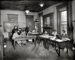 Washington, D.C., circa 1925. "Holy Name office group." An impressive array of Jazz Age office equipment and hairstyles. National Photo Co. View full size.
I see Harry Potterin Drag.
The woman on the far rightjust got a humorous email from woman third from left. LOL.
Theodore Roosevelt MemorialI believe the "Red Wheel Taxi Company" ad to the left shows an artist's rendering of John Russell Pope's Theodore Roosevelt Memorial design of 1925. This memorial was never funded by Congress and Pope went on to design the Jefferson Memorial in the same location years later. It would be interesting to find the original design of the TR Memorial and compare.
[The taxi ad shows Memorial Amphitheater at Arlington National Cemetery. - Dave]
I stand corrected. I was looking for something close to the heart of DC and should have "crossed the river" during my search before posting. Thank you for the info.
Don&#039;t want to work there!Must be a busy place, four supervisors for six clerks and a sign extolling the employees to work faster! I wonder what it was really like?
CursiveEven in so mundane a setting as that, beautiful penmanship seems to have been part and parcel of everyone's existence!  The artistic flourish in that capital "A" is worthy of framing!
HeyI think she's shopping on eBay!
Slurp.Is that a dog's water bowl on the floor to the left?
Do the Hustle!Let me get my leisure suit, disco ball and sideburns -- oops, wrong decade!
Terrific picture. It makes me thankful that I have my own desk and office!
Clothing and woodworkingMany of the photographs on Shorpy, from much earlier times, show the clothing of the period, but especially the respect that office workers, and others had for their job, etc. Another aspect of many photographs is that they show such wonderful woodworking and respective detail. More modern times, as well as current construction, exhibits a variety of detail taken from many periods in history. New ideas and designs can provide a fresh look at the use of wood and other materials.
Platen PonderingI wonder why it took so many years from the invention of the typewriter (1870) before they were manufactured with cases that enclosed the mechanicals? Certainly a case would cut down on the noise the machine made, and would prevent (especially) dirt and dust from gumming up the works.
Next I'm going to ponder why all women's shoes of the 1920s had straps. 
(The Gallery, D.C., The Office)