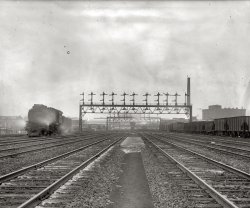 "Union Station tracks, Washington, circa 1920." National Photo Company Collection glass negative. View full size.
And the engine isThat's a Pennsy E6s on the tracks.
PRR Steam LocomotiveThe steam locomotive on the left is a Pennsylvania Railroad E class "Atlantic" type with a 4-4-2 wheel arrangement. These were the 737s of their time. They hauled short distance passenger trains at high speed. These locomotives had 80 inch driving wheels, and were capable of 100 mph.
E6s indeed ...... the size of the boiler and the delta trailing truck gives it away.  
The prototype, #5075, was built by the Pennsylvania Railroad at its Altoona shops in 1910.  This locomotive was unsuperheated (class E6).  Two superheated prototypes were built by the Pennsy in 1912, one (#1092, class E6sa) with rotary valves and smaller cylinders.  Following an extensive testing program, the PRR decided prototype #89 (class E6s) was what was wanted and built 80 duplicates in 1914.  Prototypes 5075 (now renumbered 1067) and 1092 were converted to E6s in 1913, making 83 of the class in all.  
They were reported to be the first locomotives anywhere to develop an actual (vs. theoretical) 1,000 horsepower per driving axle.  
To my knowledge, only one was preserved.  #460, the famous "Lindbergh Special" engine, is at the Railroad Museum of Pennsylvania (Strasburg PA).
(The Gallery, D.C., Natl Photo, Railroads)