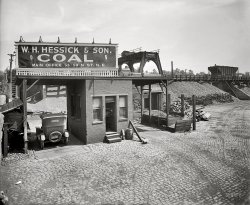 Washington, D.C., 1925. "Hessick &amp; Son Coal Co." The company's catchy slogan: "Anthracite and Bituminous Coal in All Sizes (Furnace, Stove, Egg, Chestnut, Pea) for Immediate Delivery." National Photo Co. glass negative. View full size.
Valley of ashesAll that's missing are Doctor T. J. Eckleburg's spectacles.
Two Scales...Two scales for in and out weights....Exit weight minus Entrance weight equals the weight of the coal load.
The coal came out of the bottom of the rail car into the pile below, how did they get it from the pile into the trucks for delivery? I don't see any conveyors unless they are hidden behind the building.
[Or they could just weigh the truck twice on the same scale. The coal comes out of the chute to the left. - Dave]
Black goldWhen I first looked at this photo, I immediately sensed the "smell" of coal in my mind although it's been 40-plus years ago that I last knew the aroma.
Note the worn doorway threshold; many a gritty boot has trod there. 
Clear as a bellThis is one sensational negative! Perfectly developed as well. 
True GritThat place has "Dirty Jobs" written all over it.
A coal bin in every basement....Younger people may not realize that in days of yore, the homes in cold climates (northern states) all had an area of the basement walled off from the rest of the cellar, in which a very low-to-the-ground window allowed passage of a chute (like a children's slide) with which the coal truck would deliver large amounts of coal, a ton or more, to be used by the home's occupants over the winter.  We had a coal stove and a coal-burning furnace before we switched over to oil and gas.  I would not want to go back to those laborious days.
Coal, coal, everywhere...So why is there a pile of firewood stacked to the right of the scales?
[They sold wood, too. - Dave]
The Gentle Art of Coal Delivery With regard to an earlier comment:  if you were LUCKY, coal would be delivered direct from the truck to the basement chute.  My grandfather was not so lucky:  the chute was to the rear of his house, and there was no alley.  Coal was dumped on the curb in front of his house.  He'd have to transfer the stuff around back via wheelbarrow. He was very happy to convert his boiler to natural gas.
One could still see the rail berm pictured in this photo until very recently on the block bounded by N Street to the north, M Street to the south, 1st Street to the west, and the Amtrak right-of-way to the east.  Immediately prior to the construction currently underway, the site required some environmental remediation, including the removal of underground storage tanks of some kind.
Another perspective on coalI heat my house with wood. I wish I had ready access to coal! It's hotter than wood, burns slower, and I would think cheaper too.  
Coal bins and thingsNot just in the Northern states. I grew up in North Carolina in a two-family house in the late 1940's. My father had to go to the basement to feed the stove to keep the heat up. We had ice delivery to keep the icebox chilly, but later upgraded to an electric fridge with the coils on top. For radio reception, there was a small hole in the living room floor to run an antenna wire from the console radio to the plumbing pipes in the basement to do the job.
Ah, the Good Old DaysCoal by the truckload, ice by the block, radios the size of refrigerators.  Arghhhh!  Makes you appreciate what you have today.
FossilsWe had a coal fired furnace until I was 11. Sometimes I would break up pieces to see if I could find fossils. I don't think I saw anything but plant impressions.
At one point, we had an "Iron Fireman" installed to feed coal from the bin into the furnace. I think it used an auger feed, similar to what farms use for grain.
Coal Delivery in My Old NeighborhoodIn the terraced street I lived on back in the mid-1940's, semi-attached duplexes lined one side, and garden apartment buildings lined the higher-elevation side. The coal bins for the duplexes were in the front, and chutes were used for easy delivery into the basement bins from the trucks parked on the street. On the other, apartment side, the land itself was higher, and the coal bins were in the back. Because of the lie of the land, chutes couldn't be used, so the deliverymen had to shovel the coal into very large canvas sacks and lug them up to a basement window in the rear, through which they then unloaded the coal. That must have been a job from hell.
Back-breakerMost coal trucks had tilt beds and the delivery man guided the coal onto the chute.  The guy that delivered for my dad did not have a tilt bed and actually shoveled the whole ton of coal from the bed to the chute.  I never knew what that was all about, but it was unusual.
Cold in the MorningsI grew up in Michigan, and we had a coal furnace to heat our house. One aspect no one has touched on is that no matter how much coal my dad put in the furnace before going to bed at night, it always ran out by morning.
Winter mornings it would be in the 30's or lower inside the house. I'd get dressed in bed under the blankets. There would be frost on the windows -- not on the outside but on the inside. Ah, the good old days.
Coal RenaissanceLocal antracite coal has made a comeback in Pennsylvania these days, probably due to the outrageous cost of heating oil.  One block of coal, say the size of a cinderblock, is enough to keep a woodstove hot overnight.  The stove goes in the basement below the ductwork.  Not the warmest arrangement, but a cheaper alternative.
Has the coal been watered?I remember my dad running out to ask the coal delivery driver if the coal had been watered so the coal dust didn't get all over the basement where my mother hung clothes to dry in the winter. If it hadn't been he'd have the guy sprinkle it with the garden hose. Also remember them stuffing rags around the coal room door to block the dust. Remember: "Take out the ashes!"
Dad had his first heart attack stoking the furnace on a Saturday morning. He was in the hospital for three weeks. When he got home Mom had converted the furnace to fuel oil and no more coal.
Chicago coalI'm in my mid-50s and probably among the youngest to remember coal deliveries, in Chicago (alley, dump truck, chute.) And the smell! I can't describe it. Probably for the best, but Chicago does not smell nearly as interesting as it did 45 years ago.
&quot;Them&quot; were the days!We had two coal-burning stoves in our third-story walkup. Every summer, in the heat and humidity of an Eastern big city, my mother ordered "two ton of coal" to be delivered. She claimed it was cheaper during the summer.
The delivery men had to haul up the whole two tons one burlap bag at a time. This was up rickety wooden stairs in an unlit stairwell without a handrail. I doubt they were making more than 25 cents to 50 cents an hour.
I was, at most, 7 or 8 at the time; I recall them sweating profusely. As I stood there and watched, they would pass me and still be able to crack a smile.
That was brutally hard work. They truly earned the little money they made!
Old King CoalI worked for Hessick between 1984 and 1989 and was told many times about its history in the coal business. This photo might be the old Washington Coal Depot on Rhode Island Avenue NE. I believe the coal silos are still there today.
Ashes and clinkersMy first apartment, over a carriage house on an estate, had a coal furnace that I hated. The only good part was the ashes and "clinkers" - the chunks of "stuff" that wouldn't burn. It was the best material I have ever found for putting on the ground for traction on ice and packed snow.
Coal shifting.There were conveyor belts on wheels, powered by gasoline engines, that lifted the coal from the piles on the ground into the trucks for delivery.  Deliveries to our house were made by a five-ton truck with solid rubber tires and a huge chain like a bicycle's to drive the back wheels.
It cost extra to use the chute, so my father, ever thrifty, would just have the driver dump the five tons on the sidewalk and we would shovel the coal into the basement window by hand.  It took most of the day for us to shift it into the coal bin.
In the basement, the bin was about six feet away from the furnace.  As the level of the coal was lowered, 2x8 boards were removed from the door.  
King Coal  I still use lump coal in my house and shop.  Far easier and cheaper than wood, not to mention cleaner and easier to contain.  I buy it by the barrel full.  The acrid smell is nostalgic perfume, nauseating in heavy concentration, but wonderful in small wafts.  The neighbors are all too young to know the smell and I have occasionally heard them asking one another what that "odd smell" is!  I keep it a "secret," but the DeSoto in the driveway ought to give clue that something is going on.
(The Gallery, Cars, Trucks, Buses, D.C., Natl Photo, Railroads)