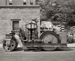 "Crawford Paving Co." Another look at that Barber steamroller circa 1925 in D.C. Watch your fingers, kids. National Photo glass negative. View full size.
Workhorse at RestMy new favorite Shorpy photo!  I love the details of the drive mechanism.  Is that some sort of clutch between the crankshaft and the worm pinion gear?
A few photos of similar beasts on the internets:

Vintage Seattle: Hi Res
Univ. Washington Library:
 [1] (note different pistons, two-speed ring gear) , [2], [3].

Nice Grease Spot, TooWhat a beast!  Just looking at it makes me want to count my fingers. 
Big WheelsI'll bet Ed "Big Daddy" Roth and Rat Fink could turn that thing into a real wheel-stander.
Ahoy matey!Did the ship's wheel come standard or is that an add-on?
Chimney linersI've not seen such a variety of chiminey liners before including the sectioned ones on the right. I wonder what is the purpose for the narrow, three section and more rectangular six section liners.  
I am thinking individual rooms surrounding the chiminey or on other floors above or below would have their own heating source individually connected to the central flue or chiminey.
That almost seems a "plumbing" pain and I am just guessing.  Perhaps it is just reinforcement.
Ring Gear  I'm scratching my head here....Where's the rest of that big ring gear?
[Behind the skirt. - Dave]
WormyThat's a pinion gear, not a worm gear.
Still (steel) around?I wonder if this is related to the Barber Green company that makes asphalt rollers today.
The fixI recall that you put your SHORPY stamp on those photos you've spent a great deal of effort restoring.  If this is true, I would like to see a before-and-after of your work with a few comments about techniques.
[This one did not take much work. A recent before-and-after example is here. Another one is below. Click to enlarge. - Dave]

ClutchMy new favorite Shorpy photo! I love the details of the drive mechanism. Is that some sort of clutch between the crankshaft and the worm pinion gear?
It appears to be a dog clutch that engages the steering wheel via the long chain. The lever behind the wheel is pushed or pulled to turn either left or right. I imagine incremental steering was done manually with the "ship's wheel."
Steamrollers for DummiesI'm pretty sure the long lever near the steering wheel is the forward/reverse control. It either allows the power to go straight down the driveshaft or then engages a planetary gear that reverses the direction of rotation for reverse. Steam vehicles don't have a clutch. They don't idle in neutral. You control the rate of speed by controlling the amount of steam allowed into the pistons. If the vehicle is stoppped the steam engine is not turning. This vehicle is why even today these things are called "steam" rollers.
Steam 101I'm no steam engineer by any means, but in all the steam vehicles that I have encountered, the engine rotation is reversed to back up. Stanleys, locomotives, ships, etc. have no reverse gear. Steam engines don't care which way they turn.
StrangeI just love Shorpy. Every time I find something new.
Why would the pinion gearset not go completely around the wheel? If that sucker ever stopped not on the gear how the heck would you get it to move again? I can't imaging anything short of a locomotive having enough torque to engage that monster!
[It does go all the way around, behind the skirt. - Dave]
Crown WheelThe large gear on the roller is a ring gear or crown wheel. We tend to call them ring gears, the Brits prefer the other. Must be sumthin' to do with the monarchy
The Barber Asphalt Company .. ..The term "steamroller" is quite correct here, as compared to "road roller," which describes internal combustion engine rollers. This Barber Asphalt Co. roller, built in Buffalo, N.Y., appears to be a very close copy of the Buffalo-Springfield steamroller. 
Barber Asphalt was not related to Barber-Greene, which never built rollers, but did build paving machines from the early 1930's, and conveyors and conveyor loaders from 1916.
Barber Asphalt was founded by one Amzi Lorenzo Barber (1843-1909), son of a Vermont Congregational preacher. After making good money in real estate, he became interested in Trinidad sheet asphalt in 1878, and founded Barber Asphalt in 1882. The company boomed with the increase in paving in the late 19th century, and by 1896 it laid nearly half the asphalt in the U.S., as well as having expanded into Europe. Barber was as good an industrialist as Ford or Rockefeller when it came to domination and lawsuits.
The first steam roller was built as early as 1865, by Aveling-Proctor in the U.K., and the last ones in the mid-1930s by Buffalo Springfield.
SteamrollerIt's not two-speed -- it was a way of making it run smoother and quieter. The inner ring teeth are in between the outer ring. The "clutch" you are talking about is the steering gear -- these rollers are direct drive. The lever at the end of the crankshaft is the reverser. 
The GovernatorAs one with warm memories of old-fashioned road equipment, I was enthralled by this photo.  The one thing I couldn't find in the shot is the part that used to fascinate me most as a kid: the spinning governor with its three balls (much like a pawnbroker sign).  The more the engine sped up, the farther the hinged arms to which the balls were fixed would swing out through centrifugal force, closing the steam feed.  With the steam feed diminished, the balls slowed, dropped, and allowed more steam in.  A remarkably ingenious but perfectly simple device.
[Those centrifugal governors have two balls. - Dave]
I yam what I yamNow, what fun would a steamroller be without a steam powered whistle? Anybody spot a whistle on board? Old cabooses used to have whistles on the rear railing for signaling. The gears, nautical wheel, and oversized parts remind me of something between Rube Goldberg and Popeye.
(The Gallery, Cars, Trucks, Buses, D.C., Natl Photo)