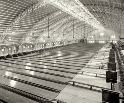 Washington, D.C., circa 1925. "Convention Hall bowling alleys, Fifth and K Sts." National Photo Company Collection glass negative. View full size.
InspirationDid the architects of the Metro stations visit this place before making their designs? 
Pin boys look out!Man, there is no protection or space for the pin boys in this design. Lots of nice maple flooring - wish I had some of it.
Pre-AMFBefore automation put about a dozen pin setters out of work.
Pin boy protectionI think the pin boys would be behind the chain link fence.  There's probably a sliding door to give them access to the "pit" behind the pins.  After the bowler rolled the ball, he could set the pins and return the ball.  This would allow each pin boy to service several alleys.  
Can you imaginethe noise in that place when it was busy !!
Northern Liberty MarketWasn't the wax museum on this site in the 1960s? Convention Hall was built as the Northern Liberty Market in 1875. It burned in the mid-1940s. Seen here previously on Shorpy. Click to enlarge.

No sliding doors for pinboysThe pinboys in old time bowling alleys had a small ledge behind the pins that they sat on, then jumped into the pit to do their work.  No chain link fences.  I bowled in places like this as a kid. Those are above ground ball returns and it looks like some sort of blackboard and chalk being used to keep score.  Also, there are no arrows on the lane in front on the foul line to aim at.  I bowled at a place in Chicago that was like that(Petersen Tournament).  After we finished bowling we would tip the pinboys by throwing coins down the alley.  Thanks for a great photo, Dave.
Pin-Spiller Paradise

Washington Post, Aug 9, 1925


Huge Alleys Will Open in Fall
Convention Hall Converted Into Large Recreation Center.

At a cost of Approximately $200,000, the second floor of the old Convention hall building on Fifth, and L streets northwest, is being converted into the finest bowling center in the United States, Canada, or any other bowling country.
It is to be thrown open to the public on September 15, and will have 50 alleys, a grandstand that will seat more than 2,500 visitors, and will cover not less than 50,000 square feet of floor space.
The work of installing the alleys is being done by experts from the Brunswick-Balke factory of Muskegon, Michigan, and is under the personal supervision of John S. Blick, former bowling champion of the District of Columbia; and president and general manager of the Convention Hall Bowling Alleys.
A decided innovation in the construction of the alleys is the shower baths and rest rooms for the women bowlers; and the shower baths and smoking rooms for the men.  In the balcony, overlooking the alleys, will be committee rooms for the league members in which meetings may be held. This will also contain a lounge for viewing the play on the vast array of alleys.  Two thousand incandescent electric lights will furnish the illumination at night. ...


Washington Post, Aug 16, 1925 


B.Y.P.U. Signs For New Alleys

The Baptist Young Peoples Union is the first organization to sign up for the use of the new Convention hall bowling alleys, was the announcement yesterday of President John S. Blick, following a meeting of the board of directors.  This organization consists of 24 teams, twelve of men and twelve of women.  Approximately 180 teams have already signed up, and many more have made applications.
...
The new alleys are to open Monday September 14, and an excellent program is being arranged which is to include a band concert, exhibitions, and souvenirs for the ladies.


Washington Post, Jan 1, 1926 


Bowlers in Finals at Convention Hall

The finals of the elimination bowling tournament in progress at the Convention Hall drives will be rolled tonight, starting at 8 o'clock. Four pin-spillers remain in the event &mdash; Al Work, Max Rosenberg, Happy Burtner and John Pappas.
A semifinal of five games will be rolled by this quartet; with the low two dropping out and the two high scorers then engaging in a five-game final for first and second place.

ContrastI'm struck by the stark contrast between that exterior and that amazing interior!  It must have really been a shock to a first time visitor to enter that building and see nothing that suggests what that wonderful exterior shell was hiding!  Marvelous.  Since it was converted from the market to this sports venue, kudos also to the unknown Architect that pulled it off.
Pin boy protection.I can now see that there is a walkway behind the fence.  I had initially seen the floor behind the fence as being the height of the ledge. But boy going in and out of multiple lanes via sliding doors would have been slow as well as dangerous since you could not see that some idiot had not thrown another ball down the alley after you crouched. I think that little raised platform every other lane is where they stand. Each ball return serves two lanes.
I have little visual experience with pin boys but all I have seen sat/stood above the lanes on a bench so they were clear unless a pin flew high.
PinsettingHubby was a pin setter in the late '50s, early '60s.  He said the pin setter would have been sitting on the flat platform on the side of the pin area.  They stepped on a lever which raised pins and set the wooden bowling pins on them (therefore, PIN boys), the bowling pins had a hole drilled in the bottom.  After setting them up, they released the pin and it dropped and the next frame could be played.  Had to be very fast.  He said if they were good enough, one boy could have done several of these lanes himself.
Had to be fastTo set up more than two lanes and get out of the way of flying pins.  The pin boys I saw in the early 50's at least had a pin placer machine that they manually filled up with the pins after each roll. They would pull the pin setter down to reset them and then would crouch in a opening between lanes untill the pins quit flying.  In my memory the pin boys only worked two active lanes at a time. 
Big ThreeToday there are three bowling alleys in D.C. Lucky Strike in Chinatown, the Hippodrome on GWU's campus, and in the White House.
AcousticsAlthough this hall would be noisy when many bowling lanes were being used, I think the height and shape of the ceiling, plus the protruding joists, would make it less noisy than more modern halls designed with low, flat ceilings.
Setters beware!I remember bowling as a kid in Michigan. The place used pin setters and because I threw the ball so hard my dad used to have to pay the setters extra to work my lane. This was in the late 50's and by then they had a rack they loaded with pins. They sat on a raised platform between the lanes and flying pins were a hazard when I bowled.
It wasn't till later in life I learned that ball revolution and angle of attack were better for scoring than speed. By then the bowling center had converted to automatic pin setters thus depriving the manual setters my new found knowledge.
That location todayFor those who are always wondering as I what those locations look like today, see this.
(The Gallery, D.C., Natl Photo, Sports)