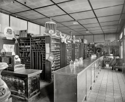 Rockville, Maryland, circa 1925. "Montgomery County Motor Co." (the Chevrolet dealer seen here). National Photo Company glass negative. View full size.
Parts Dept.This is in the smaller building to the right ("Parts," "Used Cars") in the previous post. The Seiberling All-Treads sign is in the window in both photos.
Atwater Kent RadioThis establishment was probably an Atwater Kent distributor. The company sold its radio products along with ignition parts through automobile outlets during this time. The set is a Model 24 receiver and Model M horn speaker. This is one of the few pictures I have seen showing how the set would be hooked up to a 6 volt car battery for powering the tube filaments and a high voltage dry battery for the plate voltages.
Ceiling constructionAnyone know what that ceiling is? I know drywall wasn't around in 1925 so I'm curious as to what the method was there? Some kind of plaster board perhaps with battens?
Also check out that floor. Concrete tiles?
[Drywall was indeed around in 1925. Used in the construction of many government buildings in and around  Washington during World War I.  - Dave]
Simplicity itselfJust think how simple it must have been working on those engines!  And amazingly many an old Model T got about the same mileage as today's complicated cars.
Up To DateA customer who waits in the chair had the latest Atwater Kent Radio and speaker at his command, if the batteries were up.
Not fussyIt seems to be a Chevrolet dealership that has no problem also selling Ford parts.
Have a SeatI worked for a bearing company in Fresno that started in 1912. We still had the roller bearings (like those pictured), babbit material, and chains for the drives of the old trucks and cars.  The old place had what appears to be the same style shelving, too. Would also like to note the nice chair in the right of the photograph.  Anyone think it is a Stickley??
More WeathermenSomeone's keeping serious track of the weather, by the look of those atmospheric pressure charts.
Keeping WarmAt left is the ubiquitous potbellied stove, so that staff and customer alike could keep warm while discussing that valve spring or tire patch.
(The Gallery, Cars, Trucks, Buses, Natl Photo)