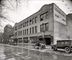 Washington, D.C., circa 1926. "Buick Motor Co. Emerson &amp; Orme garage, M Street." National Photo Company Collection glass negative. View full size.
Intakes &amp; ExhaustsI love the juxtaposition of the upright commercial brick buildings and sagging old wooden houses.  Only the building at the far left (located on the S.W. corner of 16th and M) survives today.



Washington Post, Feb 13, 1924 


Intakes &amp; Exhausts
By Si Grogan

...
Today, Emerson &amp; Orme, retail Buick dealers, celebrates their fourteenth business anniversary.  Starting in a small way this firm, composed of James Orme and Bruce Emerson, opened a salesroom and service station at 1407 H street.  They were one of the original electric car dealers in the city, handling electric commercial as well as passenger cars.  As the day of the electric car began to wane, due to the simplification of the gasoline vehicles and its wider range of operation, they added a gas car to their line.  Outgrowing their H street quarters, they moved to the present location on M street between Sixteenth and Seventeenth, where they occupy a three-story building built to suit their needs.  Shortly after moving to their present quarters they took a Buick retail car franchise and during the past few years in a the car sales department have devoted their attention to the sale of this car.  Their garage which houses the machines of many of Washington's famous people is one of the most complete and best known in this section of the country.

1620 M Street NW to-dayView Larger Map
Not big players in the art scene, apparentlyThere is a certain charm to the stark billboard and occasional Buick sign being the only cues to what the building is.  I have an idealized image of the past, where car dealerships and garages looked more like the one in this 1920 postcard:

Can anybody identify the markings near the top of the building?  I assume they represent the original owner, although I cannot identify them.
[Emerson &amp; Orme seem to have been the original owners. - Dave]
E&amp;OEmerson &amp; Orme, Washington, D.C., agents for the Detroit electric, have commenced the erection of a two-story fireproof building to contain salesrooms, service station and garage. It is located on M Street, between 16th and 17th Streets, and will have a frontage and depth of 135 ft. On the first floor there will be a salesroom, 19 by 54 ft., for new cars and a salesroom 19 by 54ft., for second-hand cars. Offices and a waiting room will be located on the main floor, together with a large  garage. The charging plant will be located on the second floor. Provision has been made for a 3rd story at some future time. The building will be completed in January. -- Automotive Industries, 1919
National GeographicNational Geographic Society headquarters now occupies the entire block with two modern buildings. The Society's original building, Hubbard Hall, still stands at the corner of 16th and M -- the corner is visible in this photo through the trees on the left.
Odd Fellows make Good NeighborsBuilding in the background with the funny dome is an Odd Fellows Hall at 1604-1606 M St NW. 
(The Gallery, Cars, Trucks, Buses, D.C., Natl Photo)