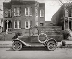 Washington circa 1926. "Semmes Motor Co. John Fisher truck." The more you look at this, the more you'll see. National Photo glass negative. View full size.
GumdropThe little girl in the window may also be wondering where the candy man is. Special delivery?
[Very good. What else do we notice? - Dave]

Daddy&#039;s HomeIs the address on the truck the same as the house? If so, that's one lucky kid. The family dentist must have made a mint off of them.
The address on the vanThe address on the van matches the number on the house? So the girl in the window is probably little Ms. Fisher.
Little Miss FisherI'd say she was the candy man's kid.
[Good. What are some other points of interest? - Dave]
PointsHe's quite protective of his spare tire.
If that thing behind the truck is fire plug, he might be parked illegally.
FireplugIs that a fire hydrant?  Never saw one like that.
[That was the first thing that drew me to this photo. -- the Mysterious Pygmy Hydranty Thing. There's a similar one here. Maybe it was some sort of shutoff valve for a water line, or a sewer cleanout. - Dave]
What?!? Daddy&#039;s HOME????As Mr. Fisher neglected to mention the midday appointment he had with the National Photo guy, at this moment there's an awkward scene taking place in a back room between Mr. Fisher, Mrs. Fisher and the mailman.
1008 Florida Avenue N.E."Capitol Hill Premium Cigars &amp; Tobacco."
View Larger Map
DetailsLooks like the truck has sliding cab windows, which must have contributed to an "interesting" entry or exit. Is the house a double? That might explain the placement of the ever-so-neatly trimmed hedge.
[Very good. The truck has a two-part door, the upper section with sliding glass. - Dave]
Hurry!I betcha he stopped by to use the can. He left the door wide open. A quick dash and back to work. I can't figure out how those windows work. Does anyone think that they're attached to the door? It's the only truck of that era that I've seen on Shorpy that has a fully enclosed cab.
[The door is in two parts, upper and lower. You would have to open each one separately. - Dave]
The lock on the spare tire.There is a chain and padlock on the spare tire, presumably to prevent someone from walking off with it.
The ThingLooking at the Google street view, it's rather distorted, but you can make out the top part of a new fire hydrant at what appears to be the same location as that pot-bellied old one. So I'm voting for that being a fire hydrant which must have had the valve on the side we can't see, because that sure isn't a valve on top of it. Unless perhaps that top flipped open and there was a valve down inside. If so, you could say for certain they don't make 'em like that anymore.
[It looks way too short to be a fire hydrant. - Dave]

HydrantIf you move one blip to the SE in the Google street view, then pivot around to the left and zoom in all the way, you can clearly see the current hydrant. I found a site that specializes in vintage hydrants (it would be foolish to think that such a thing did not exist) and sent an inquiry. We'll see if we can stump the band.
If that was a two-family dwelling, the family on the left is lucky&mdash;they have that nice little porch.
Here's the hydrant geek's reply:
That's a great photo. Is it a fire hydrant? I don't know. It is possible that it is the cover to a fire hydrant of an earlier type. If so, this would be the first documentation for this style fire hydrant in Washington, D.C., we've seen. 
Take a look at the Curran hydrant on this page. 
This style hydrant is short and is normally hidden beneath the protective cover.
In your photo there appears to be water on the sidewalk and curb near the mystery object, suggesting this is some type of water device. 
Could it be a street washer? Street washers were small hydrants used to fill water wagons, for wetting down dusty streets. It's a possibility.  
I think this is a matter for an historian or museum in DC to help sort out. Further photographic evidence, such as a hydrant in use with the same style cover sitting next to it would help.
Do let us know if you find out anything more out about this thing. I think that 1926 would be a late date for such an early covered fire hydrant, but you never know.
Either that or somebody left a pony keg on the curb.
The dark sideThe left half of the building looks darker than the right. The line goes from the hedge up to the roof. Could these have been built at separate times? Or is it a trick of the shadows?
[The bricks seem to be painted a different color on each side. Or maybe painted on just one side. - Dave]

Da DoorMany times on delivery vehicles, people would enter and exit from the right, (passenger), side. If you look closely you can see there is no sliding window on the passenger side. There probably isn't a seat on that side and maybe no door either.
The FenceComparing this against the Google street view image, it looks like the delicate iron fence in front of the property survived. This is a minor miracle.
Thanks to gcreedon for posting the Google Street View image. This is always fascinating.
The Candy TruckThe Dodge truck is approximately 1923. Dodge made a lot of commercial units at that time, basically used an open car front cowl and windshield and doors, and modified the back body to suit the use.
You can see "DB" (Dodge Brothers) on the hubcaps. The left rear fender is quite battered. Spare on the left side, with lock, and a neat fender light on the rear at the top, sort of a red and blue marker lamp.
The roadster or touring style left door has an extra sliding window attached, made with a wood frame. Presumably the driver left that affixed, and entered and exited through the curb door.
Urban ArchaeologyI can't shed any light on what the device at the curb is, but I can say that this is one of those things that keep me coming back day after day.  I love these little urban archaeological mysteries.  This post has a second feature I always find fascinating, which is a modern day comparison shot. Thanks again for the adventures.
John William Fisher Jr.Articles about Mr. Fisher in The Washington Post are rather scarce - perhaps because his business was wholesale rather than retail. The 1920 Census lists John W. Fisher, age 32, living at 1008 Florida with his brother, Edward, age 31, and sister-in-law Rosina.
From the Post, we can learn that the house was originally owned by his father: John W. Sr., who passed away in 1914 at age 49. His mother, Christine, was heir but nothing more is reported on her.  Two years later the Real Estate Transfers reveal that the brothers split ownership of the house.
Edward G. Fisher died in 1957 and merited an obituary which reveals that John W. was still living at the 1008 Florida Avenue address at this time. The confectionery business was originally started by John Fisher Sr. and both brothers worked at it after their father's death.  
As to the child in the window, Edward and Rosina had at least two children (according to his obit): Elise R. Fisher (who never married) and Edward G. Fisher Jr. (who became a Lutheran minister) - No information on their ages but they seem to have been born after the 1920 Census.
John W. Jr. died Sept 20, 1972, survived by Rosina, Elisa, Edward Jr. and 8 grandnephews.  There is no evidence of John having been married or having children.  Rosina died Feb. 29, 1980.
The Thing-SpotI see that the place where The Thing once resided now has some metal covers for what look like shutoff valves.

Dark Side of 1008 TodayThe Google Street View also shows evidence of the previously mentioned dark side/light side divisor down the bricks. I note Google has upped the resolution with their new street views. The older ones, like in San Francisco, rapidly turn to mush when you zoom in.
[I would say it's kinda scary-sharp. How long will it be before Google puts a "year" pulldown next to the zoom icon? - Dave]
Looks like a duplexThe hedge centerline seems to match the lighter/darker line, and the very bottom bricks on the left side are painted white, but not on the right side.
[It is a duplex. 1006 on the left, 1008 on the right. - Dave]
Body By FisherCan't help but thinking "Body By Fisher" would be an apt term for any customers rendered portly by overindulging in Mr. Fisher's wares.
Fire HydrantThe item in question is indeed a fire hydrant. They used to be all over the City. I do know where one is now in the Northwest section. Underneath the cap were two 2½-inch outlets with valves. 
-- Coz
[Where exactly is it? - Dave]
HydrantThe hydrant is located on the grounds of the Soldiers Home. To clarify what is under the cap. There are two male threaded outlets, each with a hand wheel valve. I have pictures of it but I don't know how to post them here. 
Jekyll &amp; HydrantMany thanks to Cosgrove for these photos of the gnomelike hydrant with a hidden personality:

I know this girl!The girl in the picture, the daughter of Edward Fisher Sr., is 89 years old. She is in fact on the way to this very location at this very moment, having just seen this photo courtesy of her niece. She is my aunt, and Ed Fisher Jr. was my dad. Next time I talk to her I'll ask about the hydrant.
Re: Aunt LizDear Aunt Liz,
It is wonderful to hear of your personal connection with this photo and to read that you were able to return to the actual site and visit with the current residents. 
Not to be too nosy, but ...

 Do you have any memories of the time when this photo was taken or of your experience living at this house?
 If you took any photos of your return visit, I am sure the Shorpy community would love to see them.
 Would you be willing to share additional thoughts or emotions on visiting the house and neighborhood now?


with gratitude and respect,
             Stanton Square

I&#039;m the Girl in the WindowI was thrilled on Friday to see a picture I had never seen before. I drove out to the house with my nephew (one of the eight grandnephews) and niece who were visiting from Vermont. I had a great visit with the present occupants. Thanks for the memories!
[Dear Liz: We are all very happy to meet you! - Dave]

Candy Man&#039;s Daughter 2009Miss Fisher, the girl in the window, returned to her old home in December 2009.
Welcome home, Aunt LizI think I speak for all of us when I say, "Wonderful to meet you!"
It continues to amaze me how we keep finding people with connections to so many of the old photos Dave shares with us each day.
MEMORIES OF 1008YES, I REMEMBER THE HYDRANT.  WAGONS CAME TO GET WATER FOR STREET CLEANING.  SOMETIMES THEY FLUSHED IT OUT INTO THE STREET.  THAT WAS FUN.
AND YES, THE HOUSE WAS A DUPLEX BUT WE DIDN'T USE THAT TERMINOLOGY IN THOSE DAYS.  WE CALLED IT SEMI-DETACHED.  I THINK OUR SIDE WAS PAINTED MORE OFTEN THAN THE OTHER SIDE.  HENCE THE DIFFERENT APPEARANCE.
FLORIDA AVE WAS A BUSY STREET.  THE OTHER SIDE OF THE AVENUE WAS OUT OF BOUNDS FOR ME BECAUSE CROSSING WAS DANGEROUS.  EARLY MORNINGS IN SUMMER THE STREET WAS BUSY WITH FARMERS WAGONS, LATER TRUCKS, HEADED FOR THE FARMERS' MARKET AT FIFTH STREET. WE USED TO GO UP TO THE GALLAUDET FARM TO GET MILK, VEGGIES IN SUMMER AND BEDDING PLANTS FOR MY MOTHER'S GARDEN.  AND MY BROTHER AND I PLAYED MAKE-BELIEVE OUT IN GALLAUDET'S OPEN FIELDS. 
I WAS BORN (IN THE UPSTAIRS FRONT BEDROOM) IN JULY 1920.  I JUST MISSED THE 1920 CENSUS! WHEN I'M 90, I'D LIKE TO VISIT AGAIN TO HAVE MY PICTURE TAKEN WITH MY THREE WHEEL WALKER TO COMPARE WITH A FAMILY PHOTO IN SUMMER 1921 OF ME IN SHIRT AND DIAPER, HOLDING ONTO THE FENCE, WITH MY TEDDY BEAR AND MY KIDDY CAR, ALSO THREE WHEELS. AND I'LL SURELY HOLD ONTO THE FENCE WHICH IS NEW BUT REPLACED ONLY RECENTLY, WE WERE TOLD.  
Thanks Aunt LizThank you Aunt Liz for your recent photo and memories of living on Florida Avenue.  I used to live a few blocks south of here and really appreciate your accounts of what the neighborhood was like.
H Street Heritage Trail!!This photo will be on a board along the H Street Heritage Trail.  The board will stand across the street so everyone will know this building's history.  The trail is scheduled to open in March of 2012. 
(The Gallery, Cars, Trucks, Buses, D.C., Natl Photo)