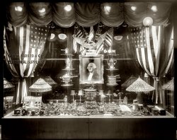 Washington, D.C., circa 1916. "George Parezo Electric Shop, 808 Ninth Street N.W." National Photo Company Collection glass negative. View full size.
Bright IdeasThis is wonderful! It strikes me as the predecessor to computer storefronts of today. I can imagine people drooling over the latest light bulbs.
The Age of ElectricityI always get the feeling that in 1916 electricity was the marvel of the age. This display has just about everything except light bulbs. There are electric motors, flashlights, irons, coffee pots, chafing dishes, a hot plate, a telegraph key(!), what look some sparking devices the purpose of which I can't figure out and some other mysterious electrical gear. And the whole thing is set off with a portrait of George Washington in (presumably) a gilt frame... lit with an electric light. In less than a decade all of this stuff would be considered commonplace and pride of place in the window would be given over to radios.
Spark GapsThose "sparking devices" are likely spark gaps used in amateur radio transmitters of that era. The advent of WW1 caused the shutdown of that aspect of radio for the duration (and if the US Navy had had its way it would never have returned). There is also a nice collection of knife switches on display. The "rocks" in the center are probably chunks of galena used (in much smaller sizes) as detectors in simple radio receivers of the era.
Tubular RheostatWell, since there is a flurry to identify widgets in the window - I'll pick out the item at the very extreme right.   It is a Wheatstone tubular rheostat (i.e. variable resistor).  The backrooms of the physics labs when I went to college were full of these things.  The physics department at Kenyon College currently has an excellent page on historical rheostats and resistance boxes with images of similar devices.
Another curiosity is associated with the language in the display for Dim-A-Lite. I'm tickled by the advertising line that Dim-A-Lite "Saves Current."  These days advertisers would proclaim "Saves Energy." Both statements are correct, I just don't think many people today would immediately identify with the concept, or need, to "save current."  
tterrace has already commented on this gadget in the previously seen image of Mr. Parezo's store.   Typical of many things which appear on Shorpy,  a google search for "Dim-A-Light" currently brings up a link to Shorpy in the first page.    
TimelySomehow, the Fourth of July springs to mind.  Not sure why, but there's something about this particular picture!
(The Gallery, D.C., Natl Photo, Stores & Markets)
