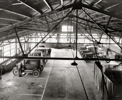 Takoma Park, Maryland, circa 1928. "Hendrick Motor Co. garage." Our third look at this establishment. National Photo Company glass negative. View full size.
High PointGreat picture, great angle.  
Early Signs of SafetyThere are two encouraging safety developments apparent in this photo (versus many other early machine shops).  The M-G set at the back of the shop has its belt guarded.  And the oxyacetylene torch rig next to it appears to have the cylinders secured to the cart.
Hendrick gives a darn about his employees!
Slow design developmentIt's interesting that in 1928 they were still more or less using the old horse carriage design.
Hold the grease, pleaseI love the details.  I see what look like protective covers over the steering wheels and seats of the cars that are in for service.  I guess greasy handprints have always been a hazard of taking your car to the mechanic.
The vise squad.There's an old saying in the trade that when building a repair shop you start with a good vise and build the shop around it.The humble vise hasn't changed in hundreds of years and in this high tech age the modern shop still can't fuction without it.
Sultry winchesI was rather surprised to see metal roof trusses in that era. I was even more surprised to see a stout wooden beam spanning two of them with a travelling block and tackle hoist running along it.  That seems like a recipe for disaster - especially when the load shifts to either side and onto a single truss.
Service, please!As a veteran mechanic, I remember the days when a valve grind was necessary every 60,000 miles. Now if you don't get at least 200K on an engine before it requires major work, you haven't maintained it correctly. The demise of carburetors and the improvement in the quality of gas and oil has reduced the amount of carbon produced in the combustion chamber, which held the valves open slightly, causing them to burn. It was not uncommon to do a valve grind and re-ring at 30,000 miles back when these cars had a few years on them.
What a neat garage!The first row of cars on the left show a Model T two-door, about 1923, then about a 1923 coupe, then another two-door, 1926 or 1927, and a roadster pickup at the end.
On the right is the best part of the photo. A 1927 roadster, and a 1927 Ford coach or tudor. They could be still-new cars for sale. 
The closest car on the right is the "New" 1928 Ford. This is a commercial chassis, probably a ton by the size of the springs, with no body attached. On the commercial cars the radiator and headlamps were painted black. Passenger cars had nickel plating. You can see the spare rim mounted in the middle.
In front is the wash bay, for excellent service.
Cold, hard workIt's sort of romantic to others who have not worked on cars, however, you might consider what it might be like for you to pick up cold spanners on a winter's morning, graze your knuckles, bend your back to lift a cast iron cylinder head off a block and after a few years perhaps suffer a prolapsed disc in your back from all the bending over car engines and then have to find other work.
Hard slog in a place like this.
(The Gallery, Cars, Trucks, Buses, Natl Photo)