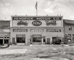 Takoma Park, Maryland, circa 1928. "Hendrick Motor Co., Carroll Avenue." National Photo Company Collection glass negative. View full size.
Fordor SedanThe new Model A Fords must have been painted on the building during the period that Ford was transitioning from the seriously outdated Model T, to the all new Model A. The Tudor Sedan shows a fresh air vent in the lower cowl. No production Model A ever had such a vent.
Legible signsI am struck by the bold, clear and legible use of typography in these older photographs. This is in distinction to the blur of pictograms, logos, and hen scratching that passes for public communications nowadays. Just look at the ads all around us. There was something deeply confident as well as respectful of potential customers embodied in our forefathers' use of graphics.
Model T SportscarAn interesting picture. I'd like to take a spin in a car featured on the sign, that Model T Coupe with the rumble seat.
[That's a Model A roadster. - Dave]
Radio DaysThe sign next to the service entrance says "batteries delivered to your home." That's because the majority of home radios in 1928 required a 6-volt lead acid battery to supply their tube filaments. Radios that ran on house current were just coming to market in 1928.
Takoma FordThis dealership later became Takoma Ford. Our family bought several cars from them because the service manager was a neighbor.  The mechanical service area was through the big opening in the picture.  To enter the body shop underneath you had to drive around the block.
It&#039;s a Sport CoupeThat would be a sport coupe... identified by the landau bars.  The top does not fold down as it does on a roadster.  Roadsters also did not have roll-up windows.  
ObservationsObligatory "you can see the photographer in the reflection."
I wonder if this was a planned photo. You'd think they'd make an effort to clean up the lot.
Also, I'm surprised the barber shop didn't make it with all of the men having time to kill while their cars were being repaired.
Fordor ventsThe car on the right side of the sign is a Fordor rather than a Tudor. The early 1928 Fordors did indeed have cowl vents but they were eliminated shortly after introduction. I have a '28 leatherback Fordor like that.
Early 1928 SedanThe early 1928's did have a cowl vent on the lower driver's
cowl. These were called the "AR" model. The early cars had a number of changes in them, as discussed in the book "Henry's Lady" by Ray Miller. So it's an early 28. Thanks.
Cheap GasThe "Gasoline at D.C. Prices" sign is humorous--today it seems like gas gets cheaper the farther you get from urban areas (at least here in the NY/NJ/PA tri-state area); apparently that wasn't always the case.
I wonder if the founders of this dealership are any relation to the Hendrick family in North Carolina that runs several mega dealerships--and a rather successful NASCAR race team.    
(The Gallery, Gas Stations, Natl Photo)