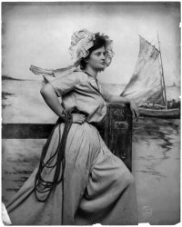 A woman wearing bonnet poses on a photo set of a pier with a painting of a fishing boat in the background. Photograph by Fritz W. Guerin, c. 1902. View full size.