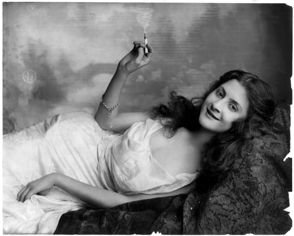 A young woman reclines on a sofa holding a burning cigarette. Photo by Fritz W. Guerin, c. 1902. View full size.