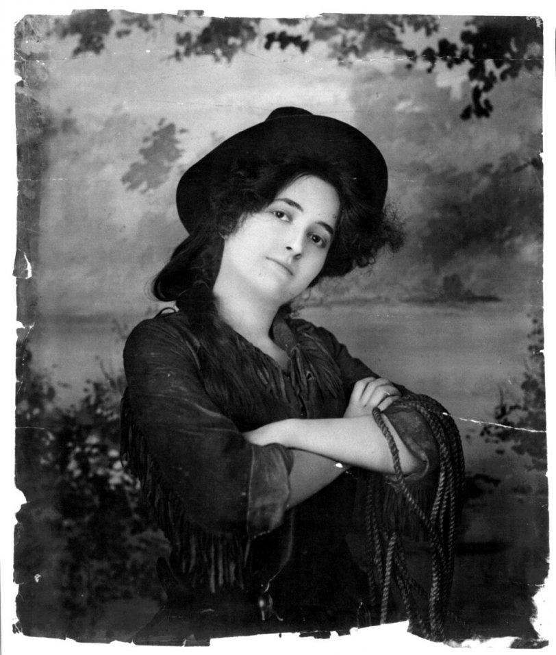 A young woman models, wearing a cowgirl outfit and holding a rope. Photo by Fitz W. Guerin, c. 1902. View full size.