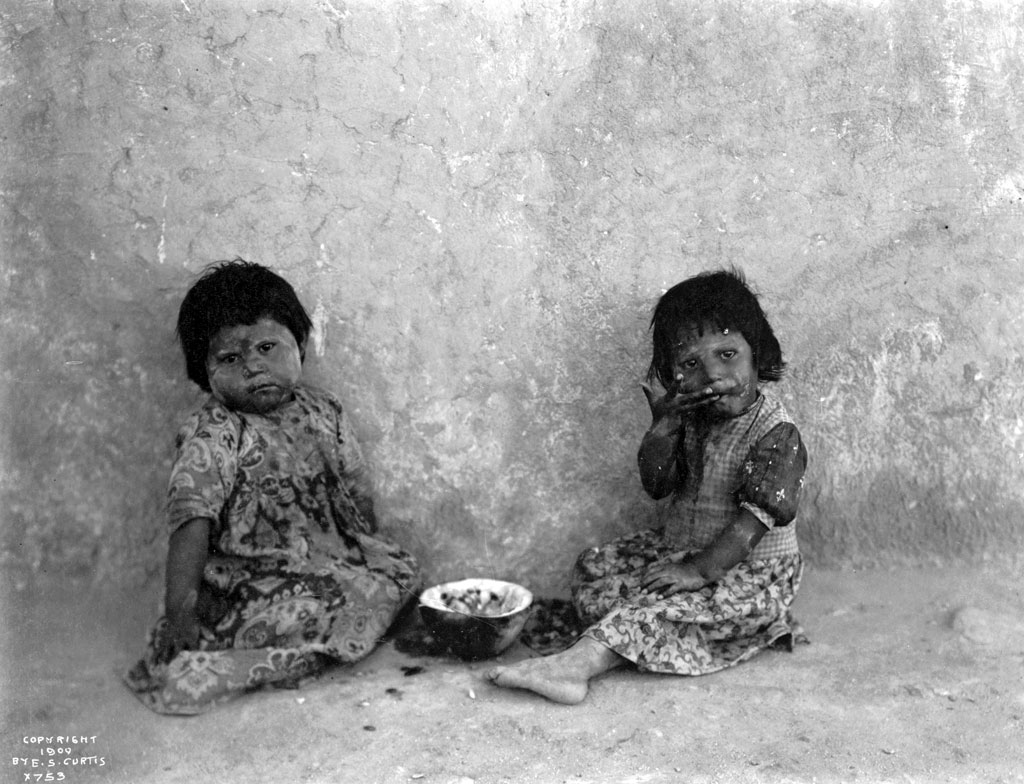 Two Hopi girls sit on the ground eating melons. Photograph by Edward S. Curtis, c. 1900. View full size.