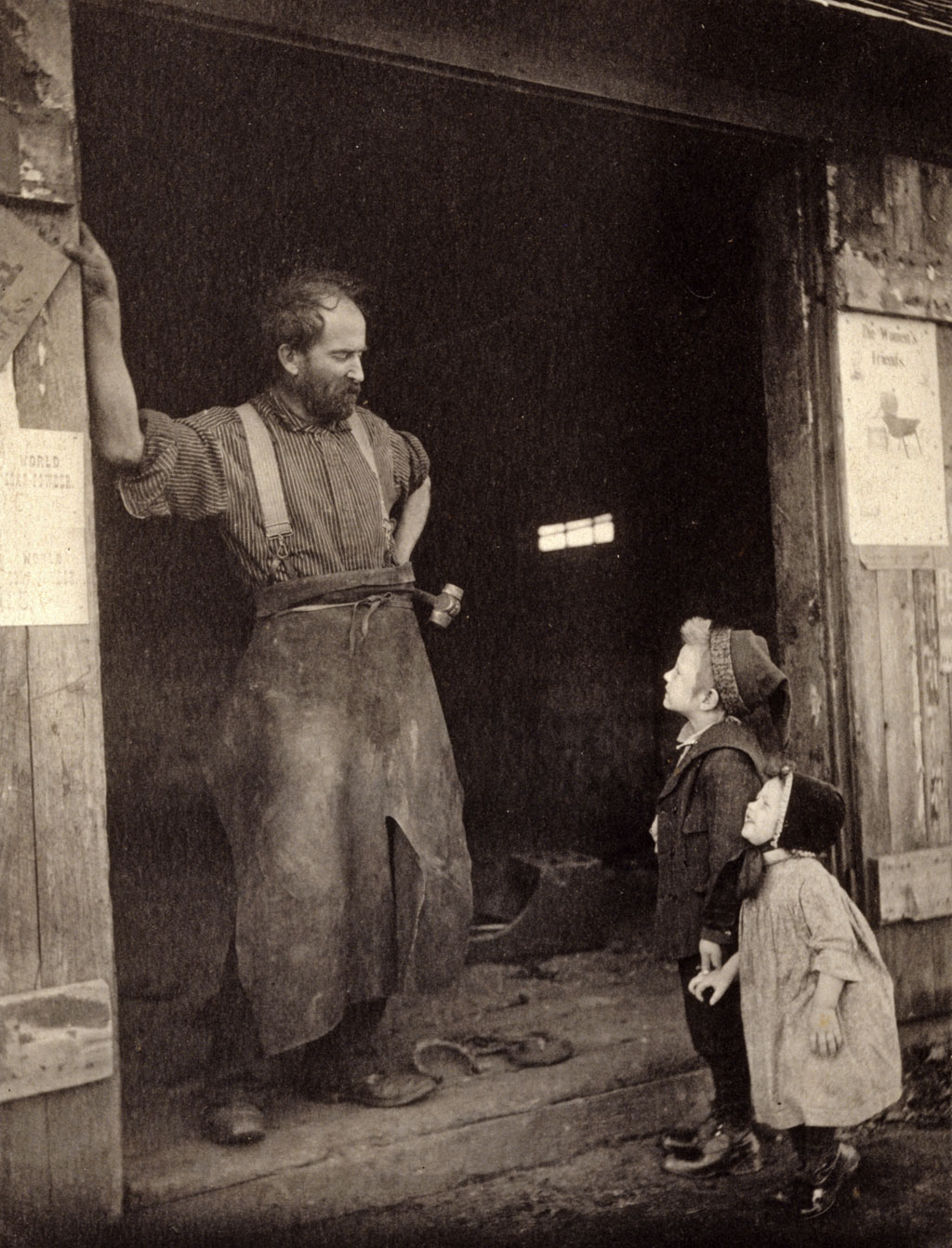 Two children talk to a blacksmith standing in the doorway of his forge. Photograph by Frances S. Allen, c. 1900. View full size.